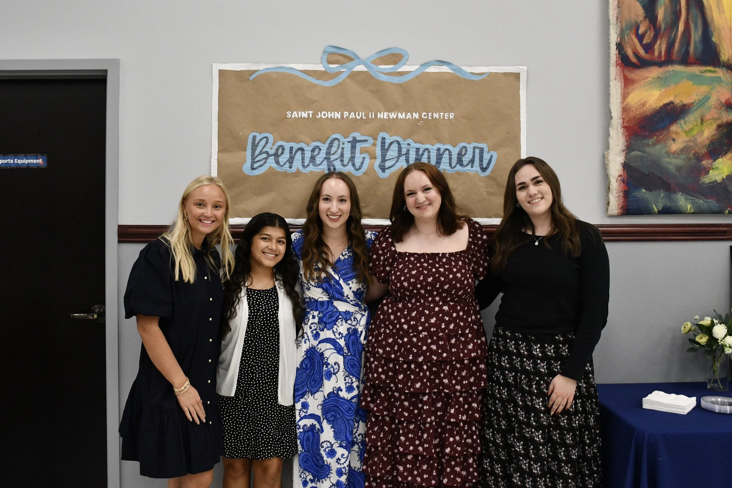 Group of five young women standing together at a benefit dinner event, smiling in front of a sign that reads "Saint John Paul II Newman Center Benefit Dinner," with colorful artwork and a table with white napkins and flowers nearby.