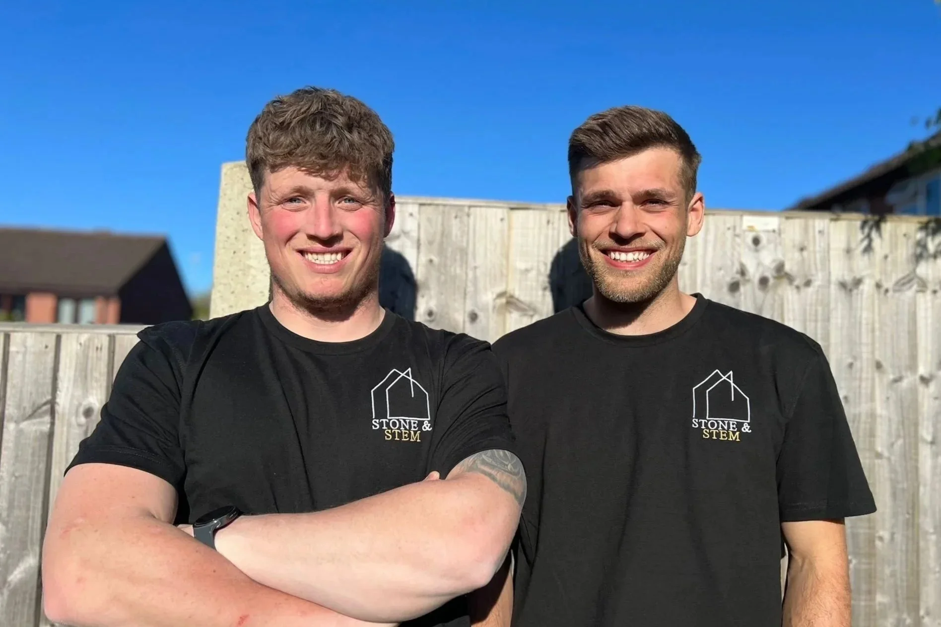 Two smiling men standing outside near a wooden fence, wearing black t-shirts with a logo that says "Stone & STEM." One has curly light brown hair, the other has short dark hair. Both are smiling at the camera.