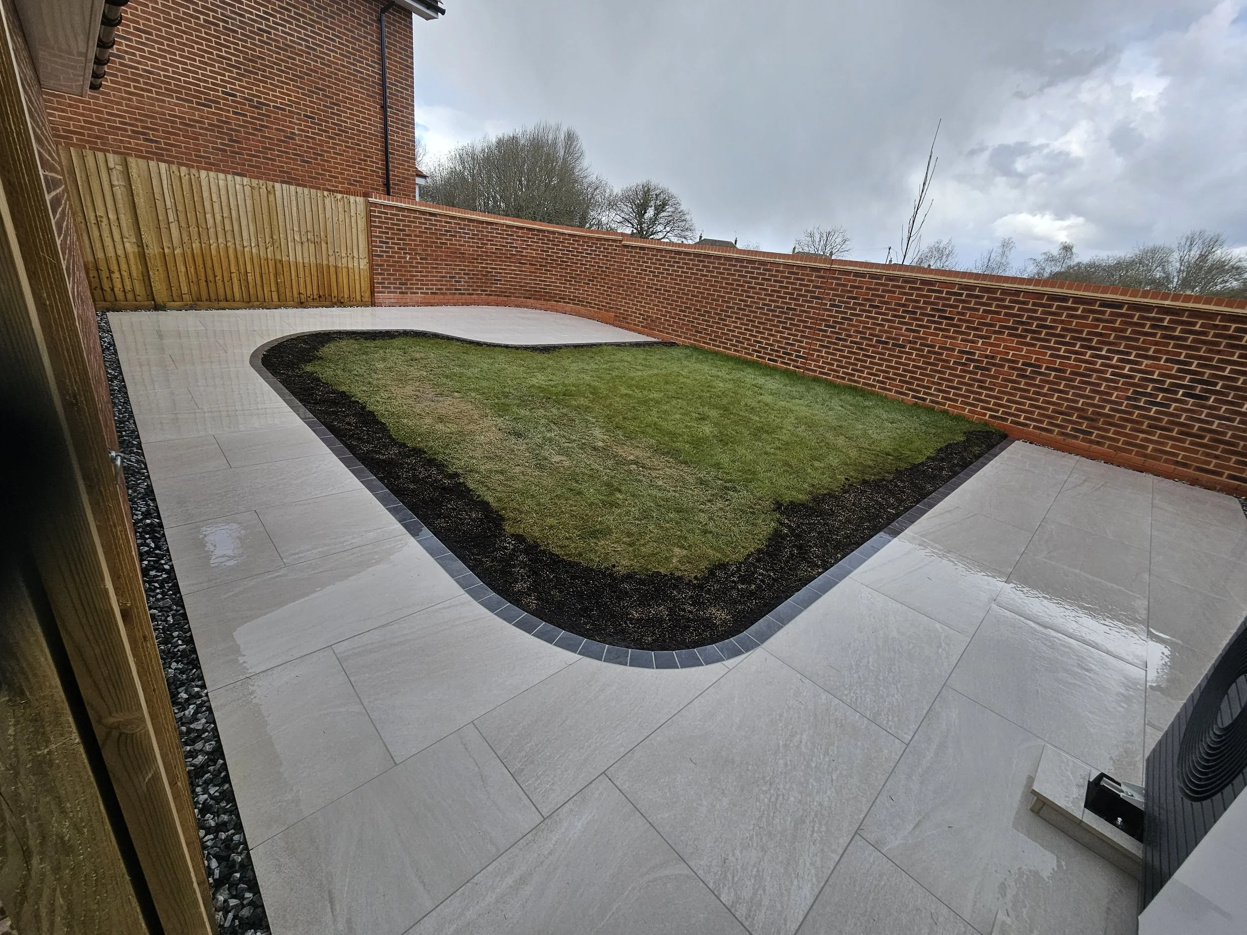 Backyard patio with a green lawn, white tiled walkway with black borders, brick and wooden fencing, and cloudy sky.