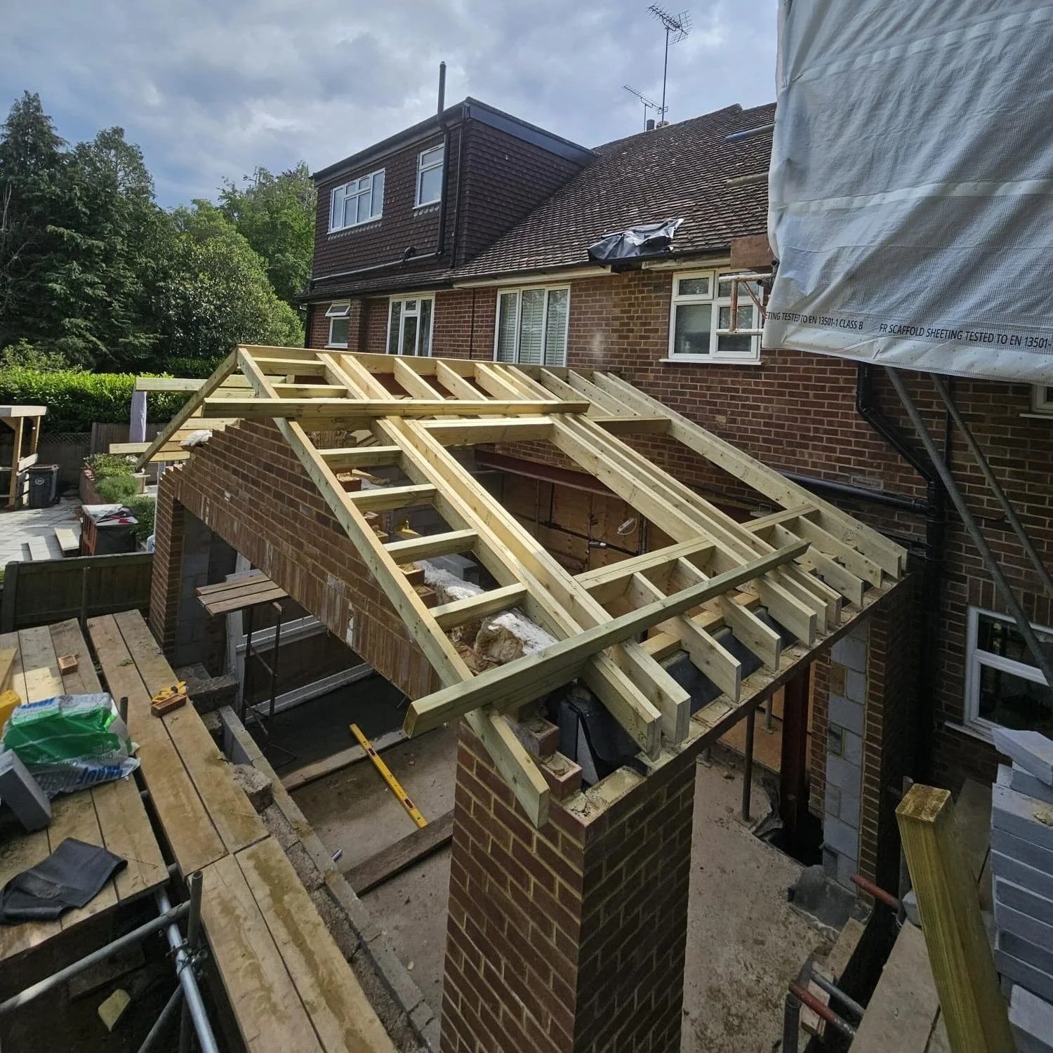 Construction site of a house roof with wooden framing, brick chimney, and scaffolding, near a brick house with multiple windows, trees in the background, and construction materials in the foreground.