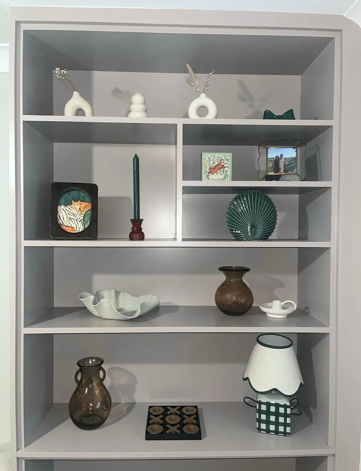 White bookshelf with decorative vases, framed photos, a green fan-shaped dish, a large brown vase, a white abstract bowl, a lampshade, and a small tic-tac-toe game.