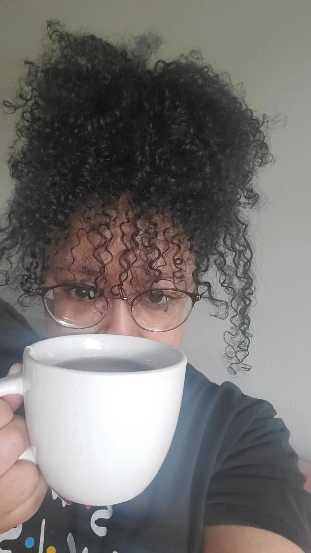 Person with curly hair and glasses holding a white mug filled with a dark beverage.