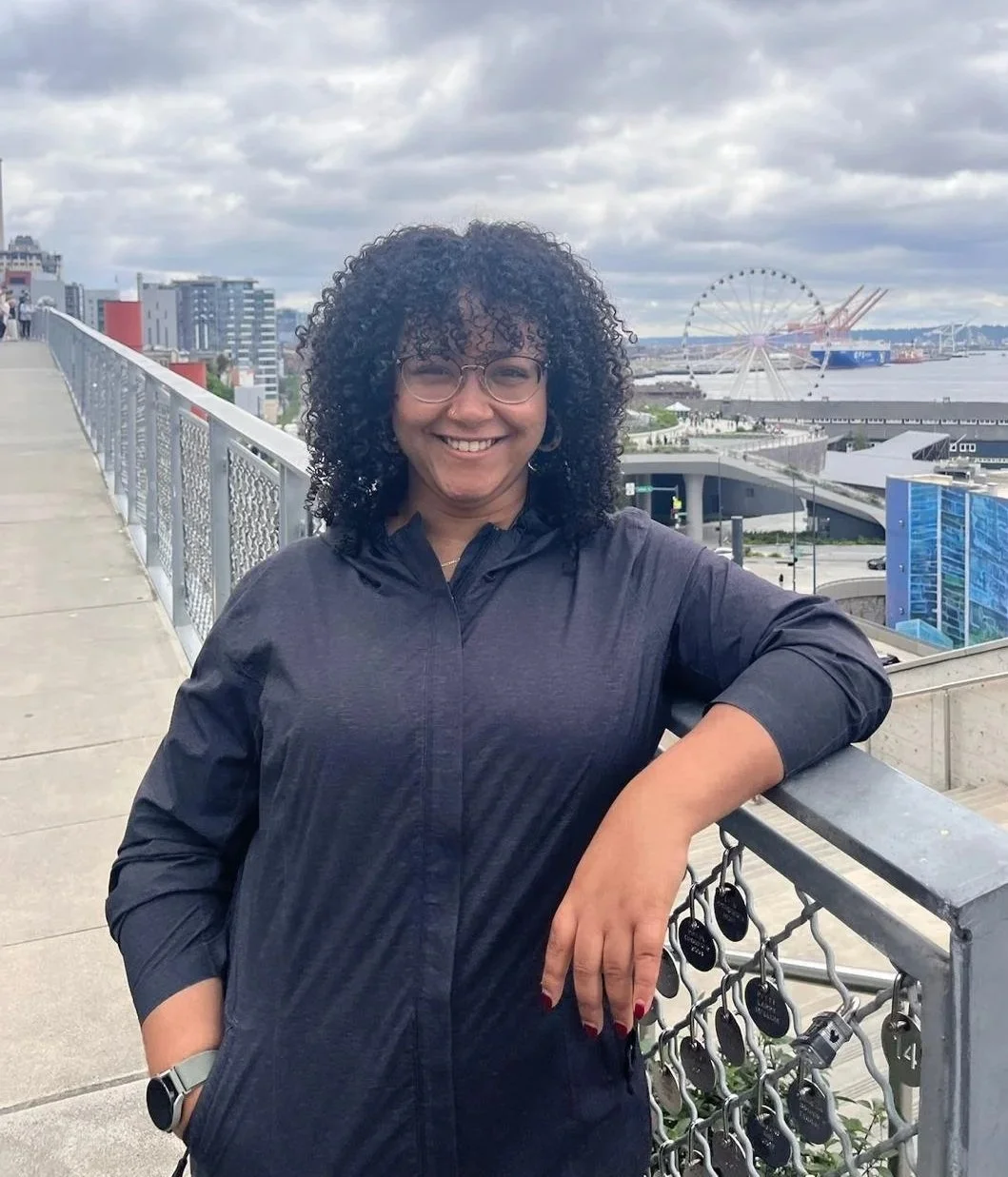 A woman with curly hair, glasses, and a black jacket, smiling on a Seattle city riverwalk with a Ferris wheel and modern buildings in the background.