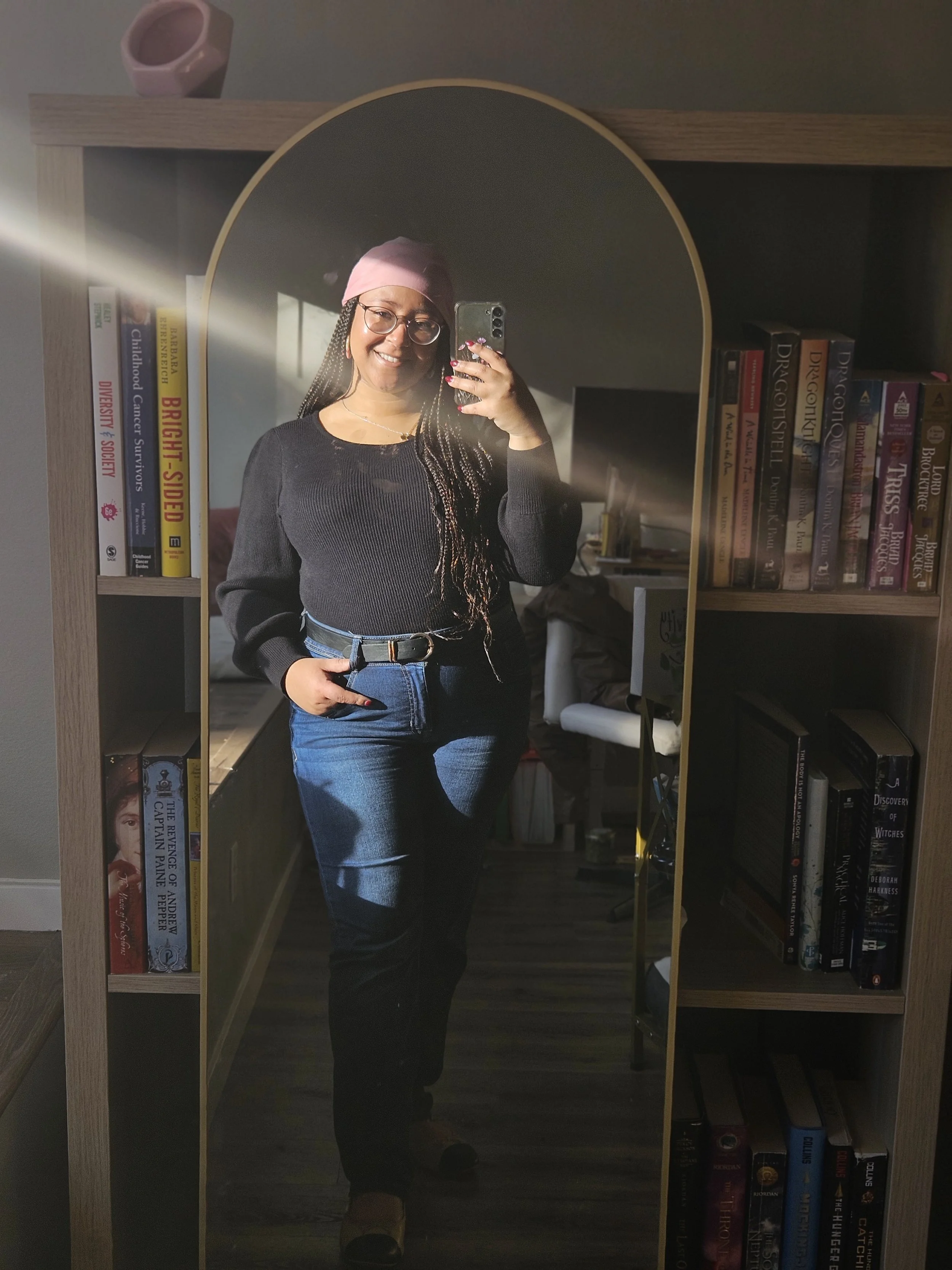 A woman taking a selfie in a mirror, wearing glasses, a pink headscarf, a black sweater, and jeans, standing in front of a bookshelf filled with books.