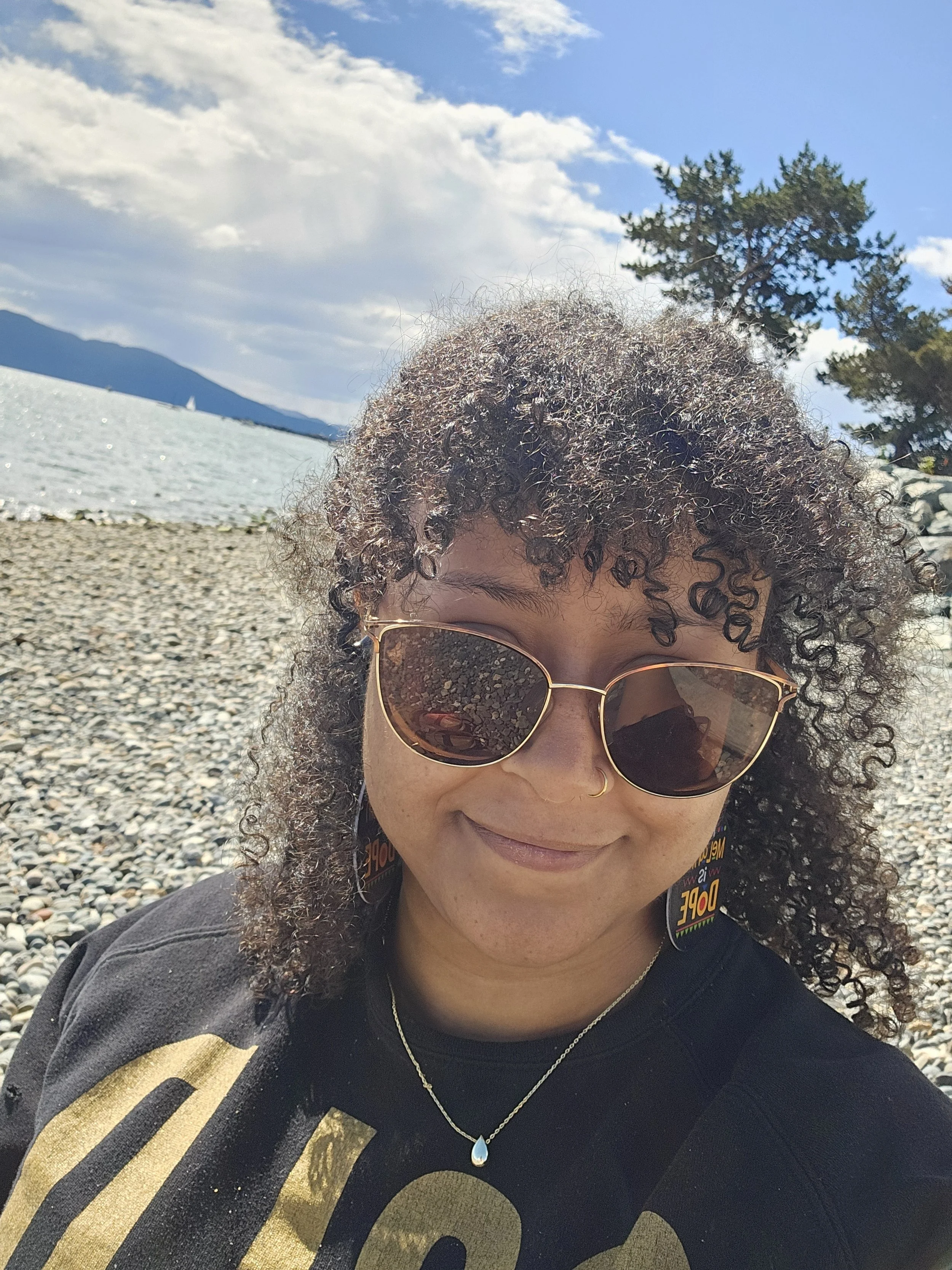 A woman with curly hair and sunglasses taking a selfie outdoors near a beach with mountains and water in the background, wearing a black shirt and a silver necklace.