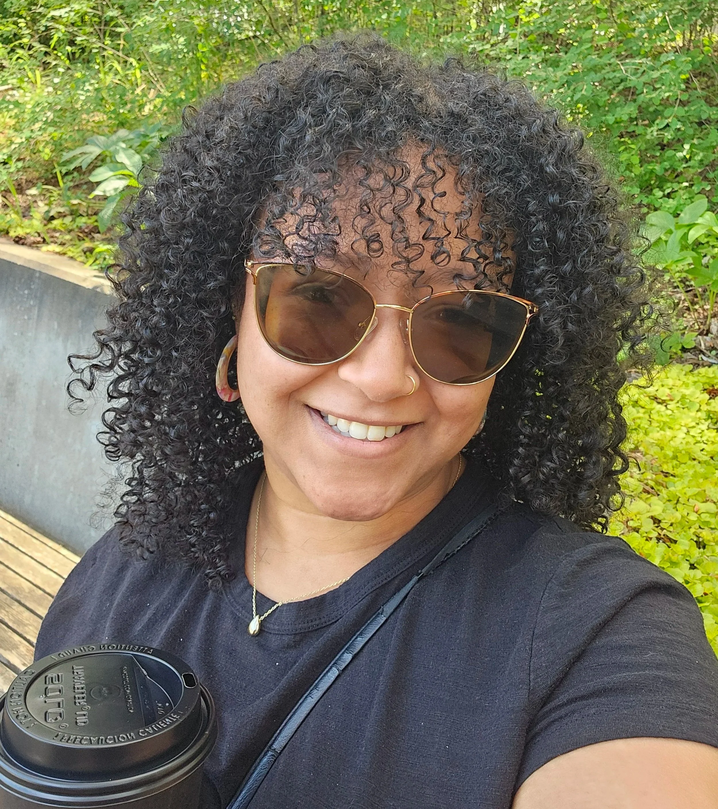 A woman with curly black hair wearing sunglasses and layered necklaces, smiling outdoors. She has a camera strap around her neck and is holding a camera with a large lens. The background features green foliage and a wooden deck.