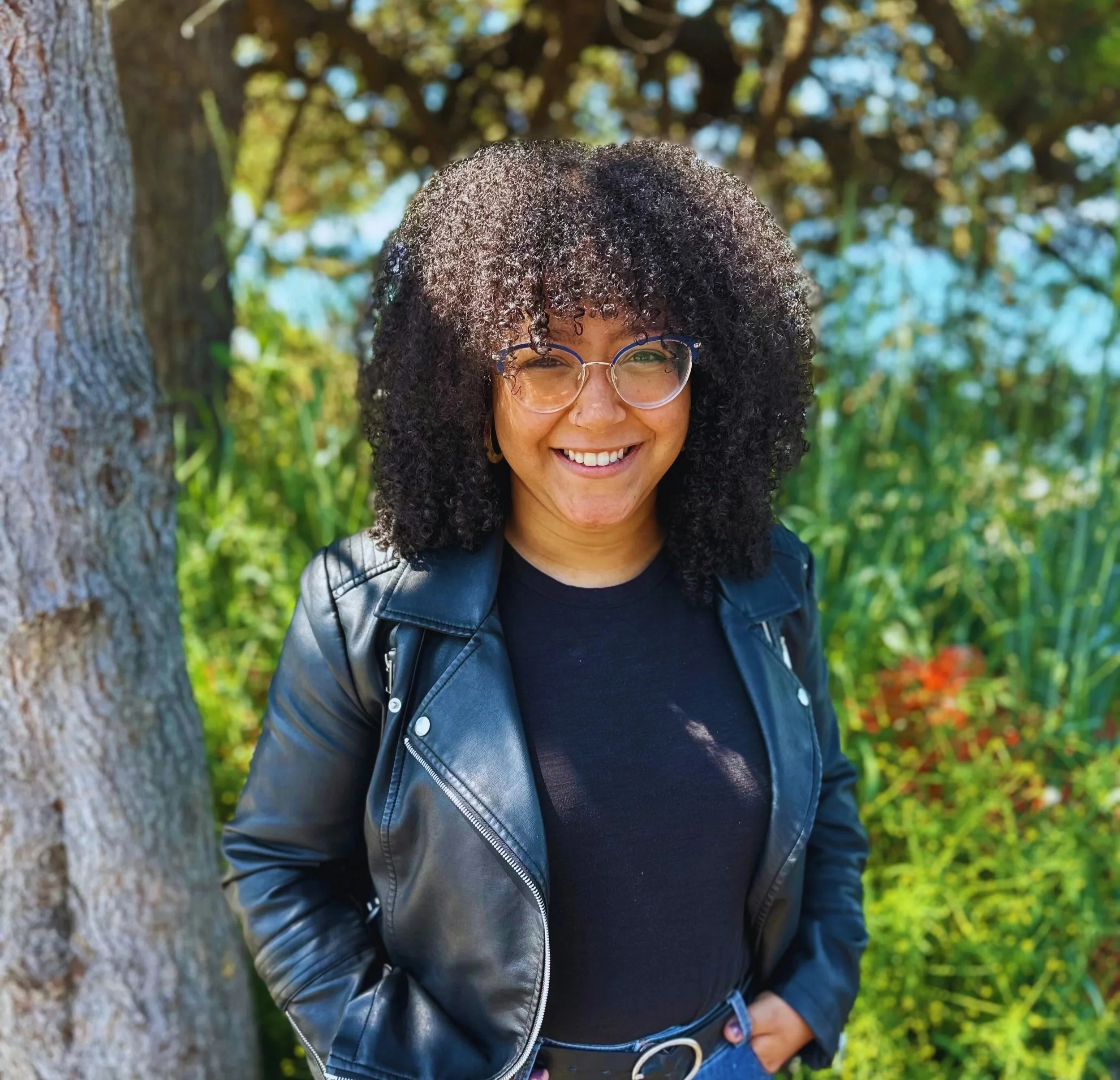 Woman with dark curly hair, glasses, and a leather jacket standing outdoors near a tree, smiling in a green park with sunlight filtering through the leaves.