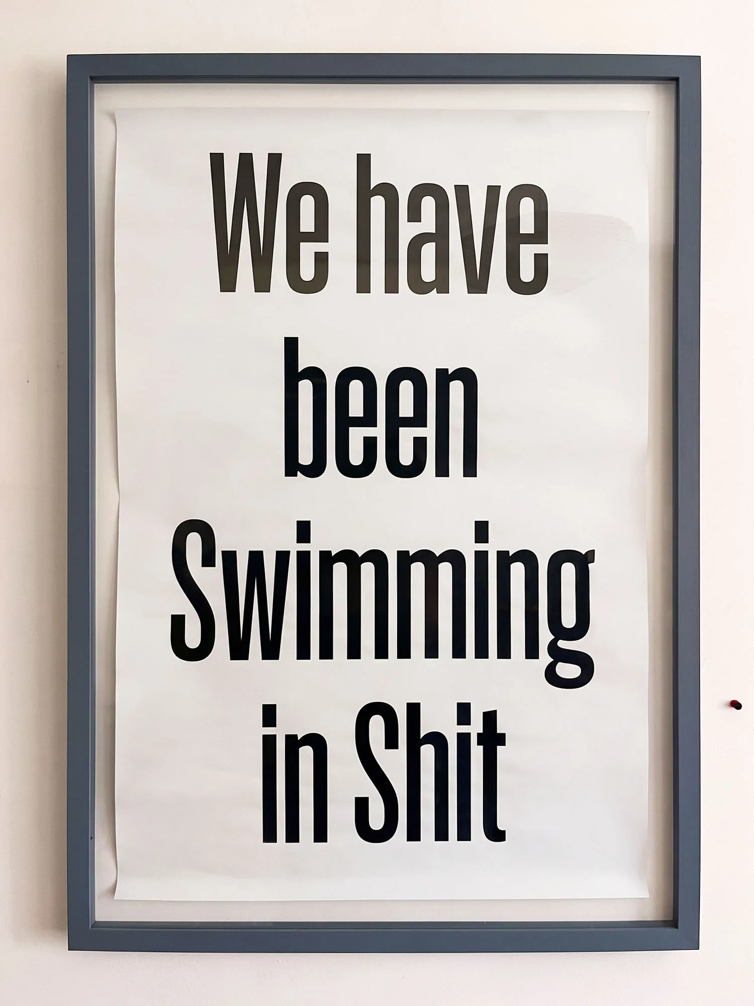 A framed Jeremy Deller poster with the text "We have been Swimming in Shit" printed in large black letters on a white background. The frame is light blue.