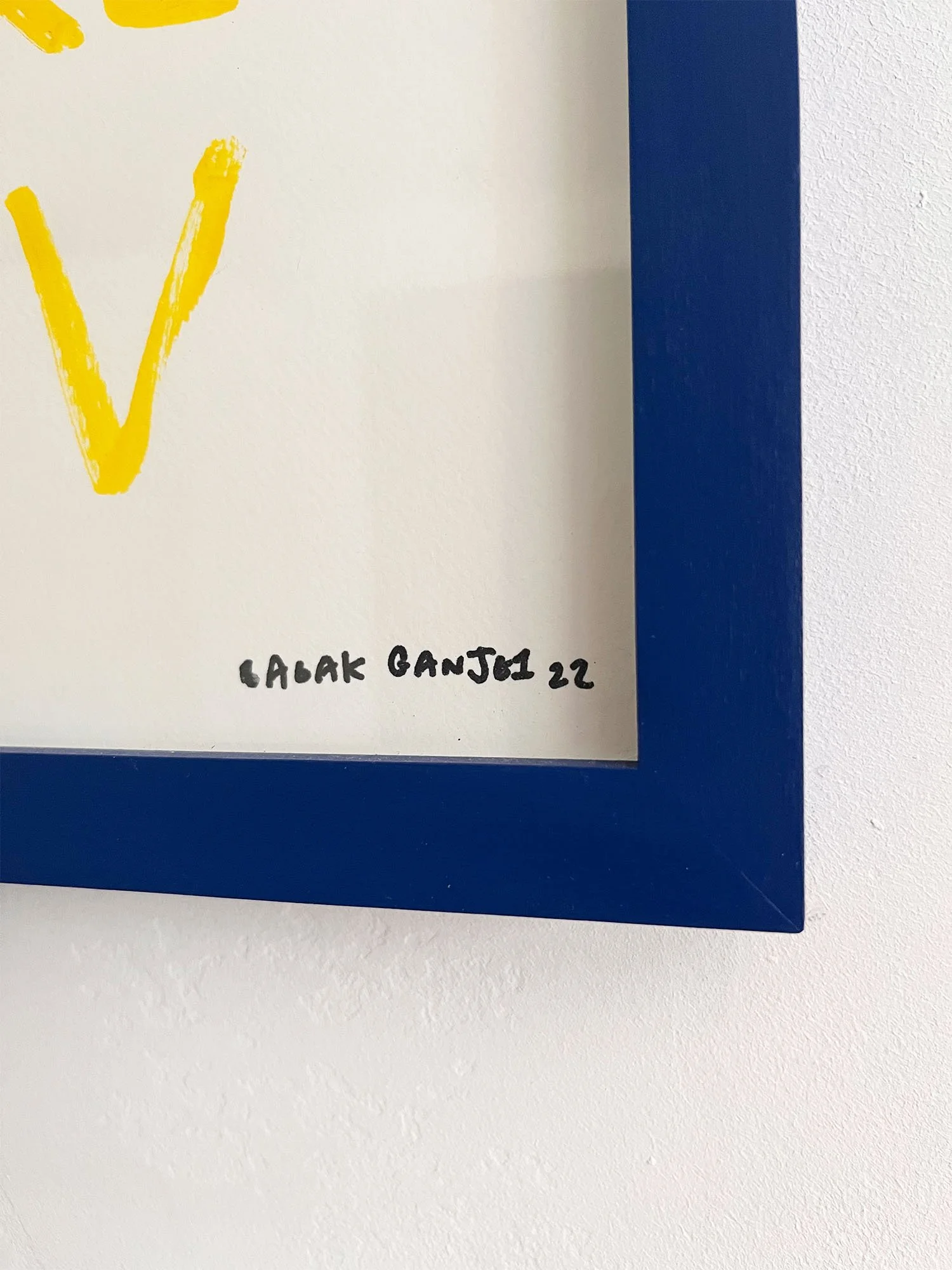 Close-up of a framed hand written print by Babak Ganjei, blue frame with white print, handwritten signature in black and a glimpse of the yellow text of the artwork.