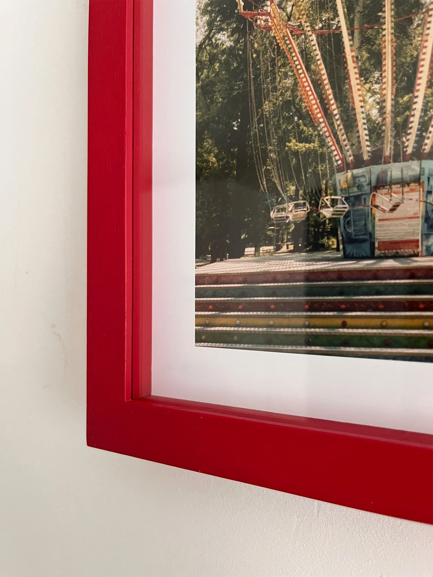 Corner detail of a framed photograph of a carnival swing ride by Venetia Scott, framed in a red frame, on a white wall.