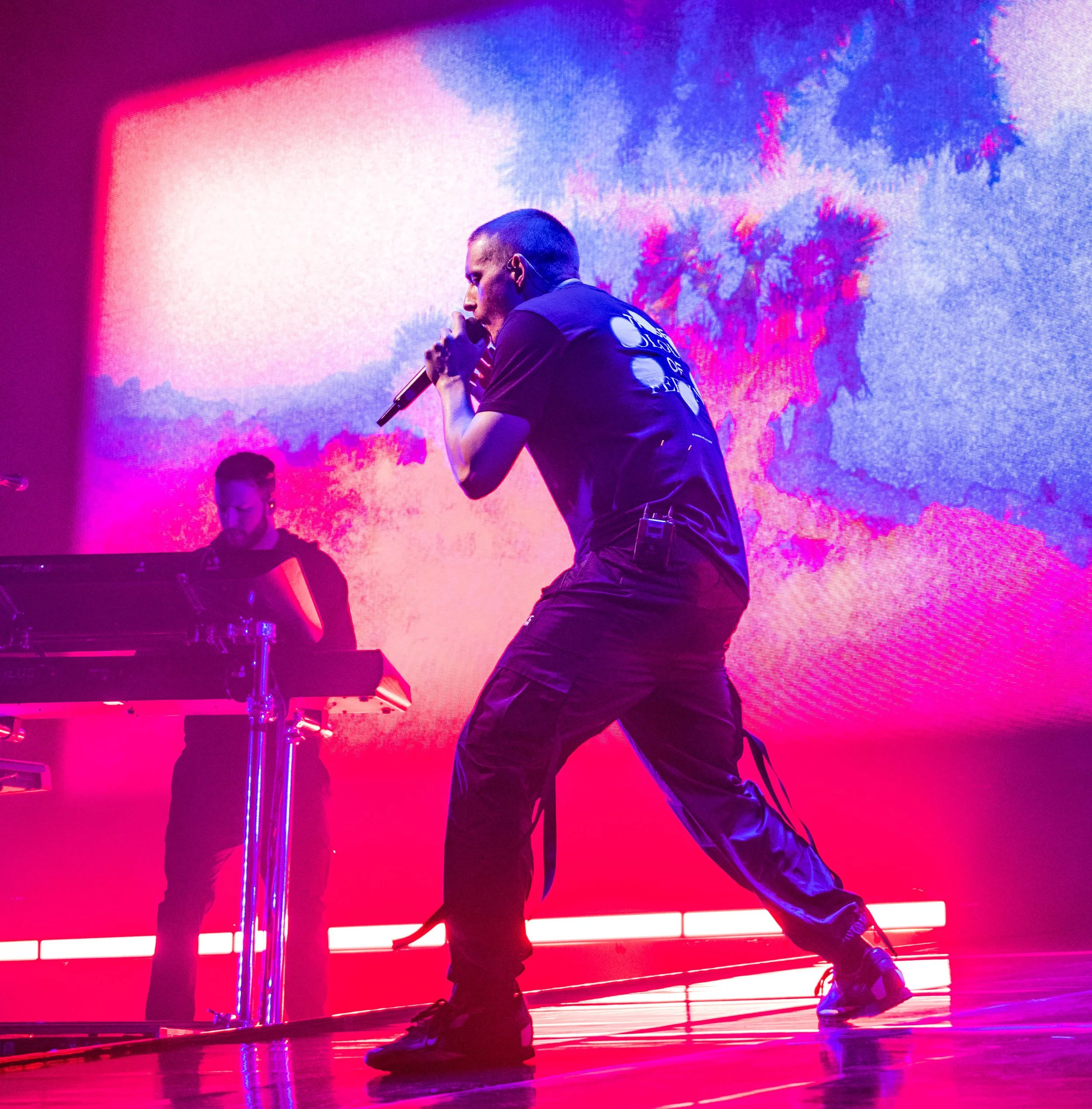 A man singing into a microphone on stage with a colorful led backdrop displaying vibrant pink, purple, and blue hues.