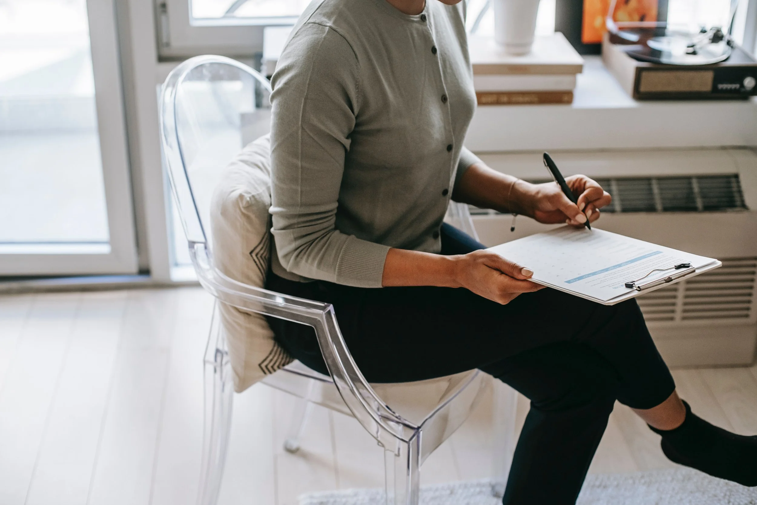 Person sitting on a clear plastic chair, holding a clipboard and pen, working at a desk near a window.