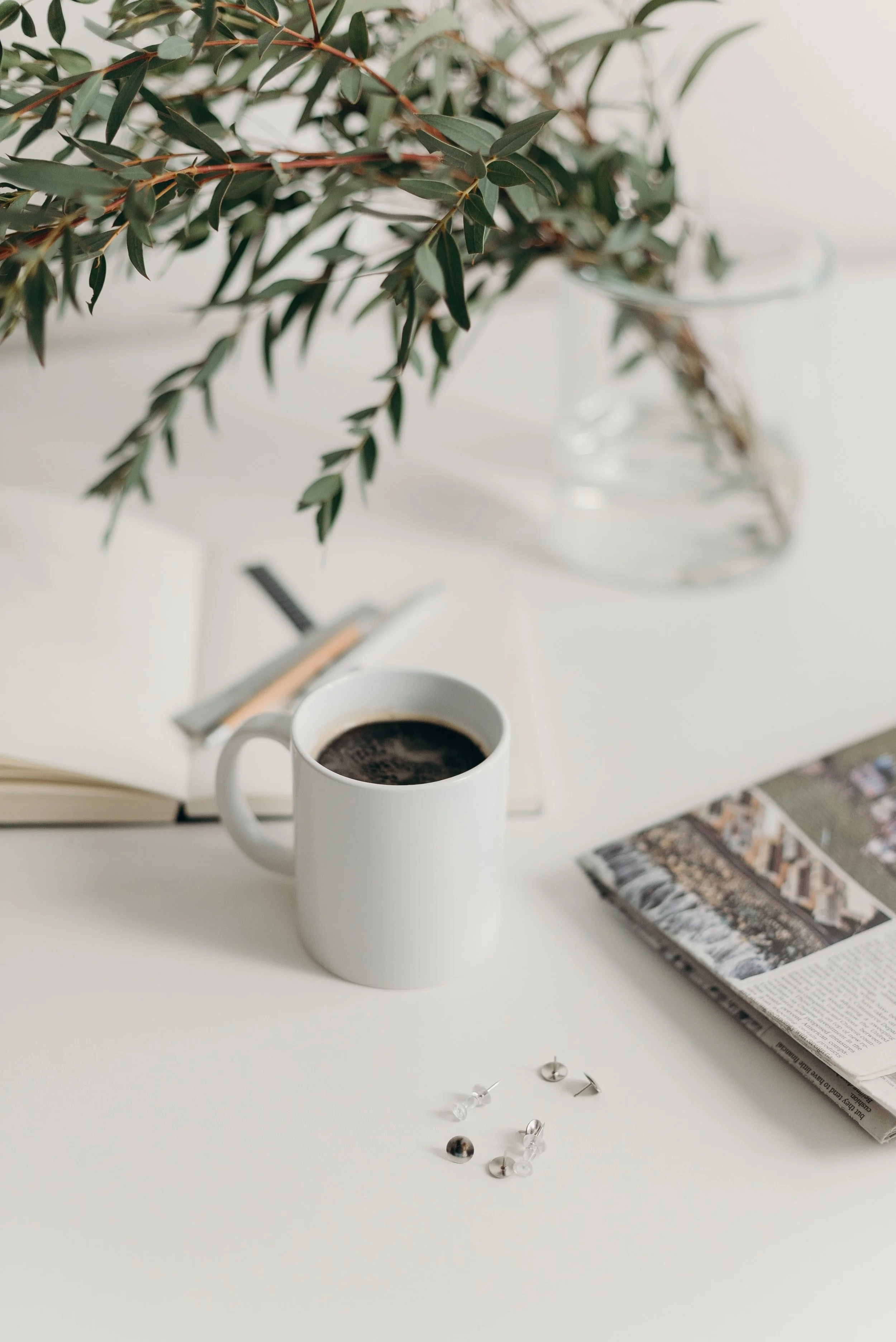A white cup of black coffee on a white table with scattered push pins, a remote control, a magazine, and a pair of silver earrings, with a plant in a glass vase in the background.