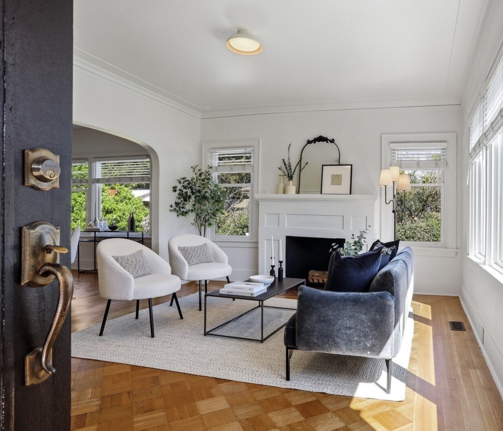 A living room with white walls, hardwood flooring, a fireplace with framed art and vases above it, large windows with blinds, a black coffee table with books and decorative items, white and dark gray armchairs, a beige area rug, and potted plants.