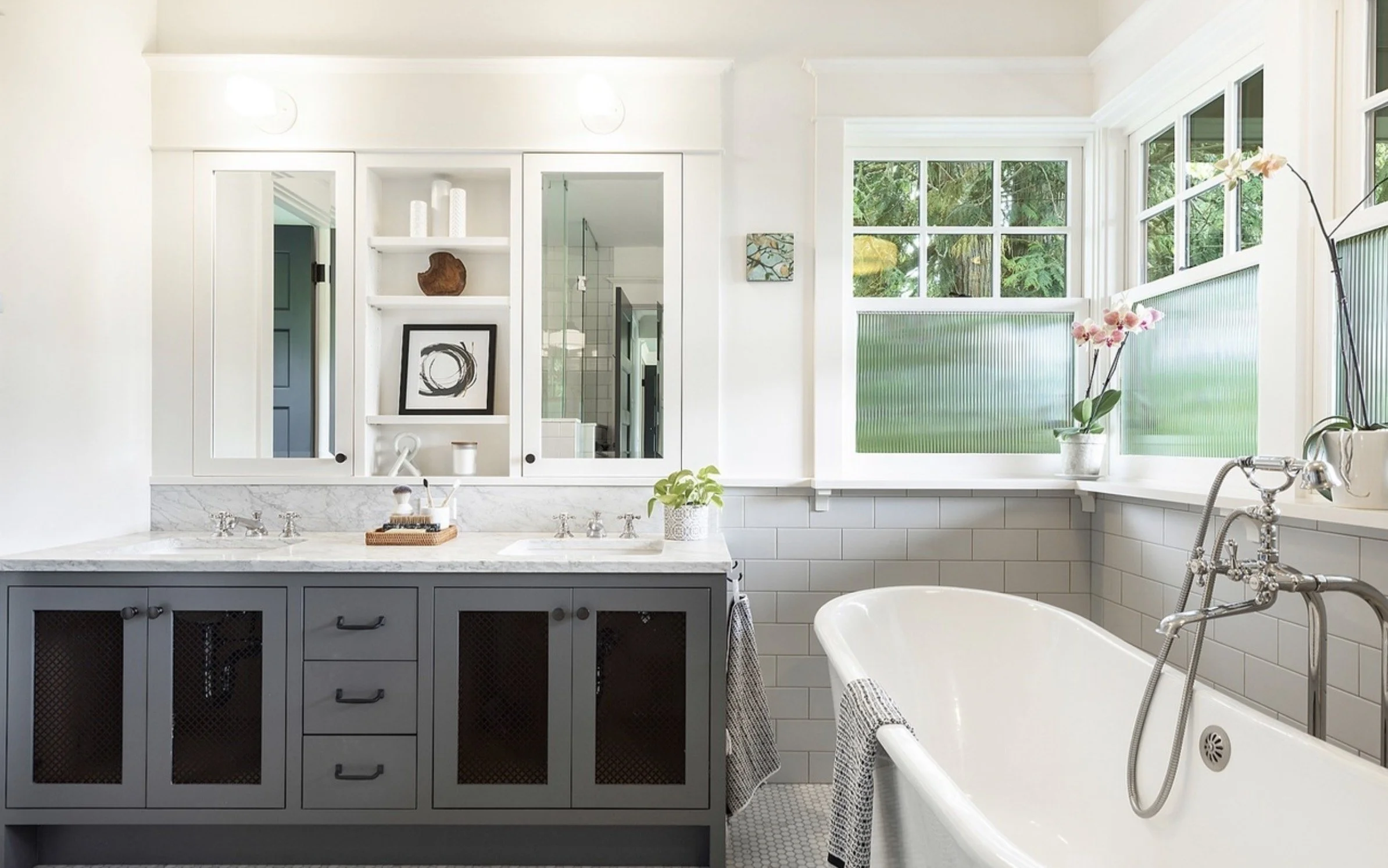 A bathroom with white walls and large windows showing green trees outside. There is a white bathtub with a vintage-style fixture and a dark gray vanity with a white marble countertop, a framed abstract art piece, and decorative items on open shelves.