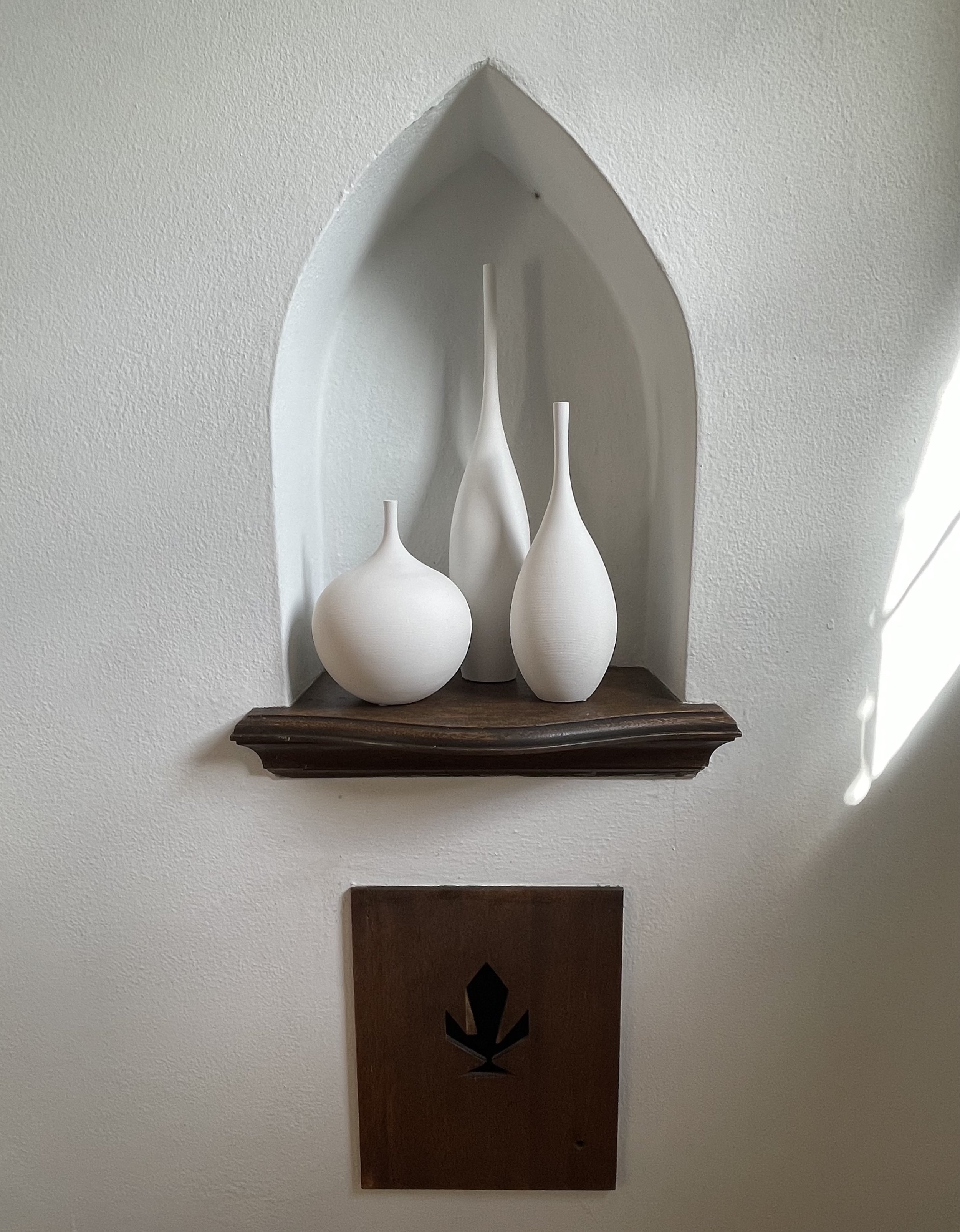Decorative wall shelf with three white ceramic vases of different heights, set within a pointed arch wall niche, with a wooden base below.