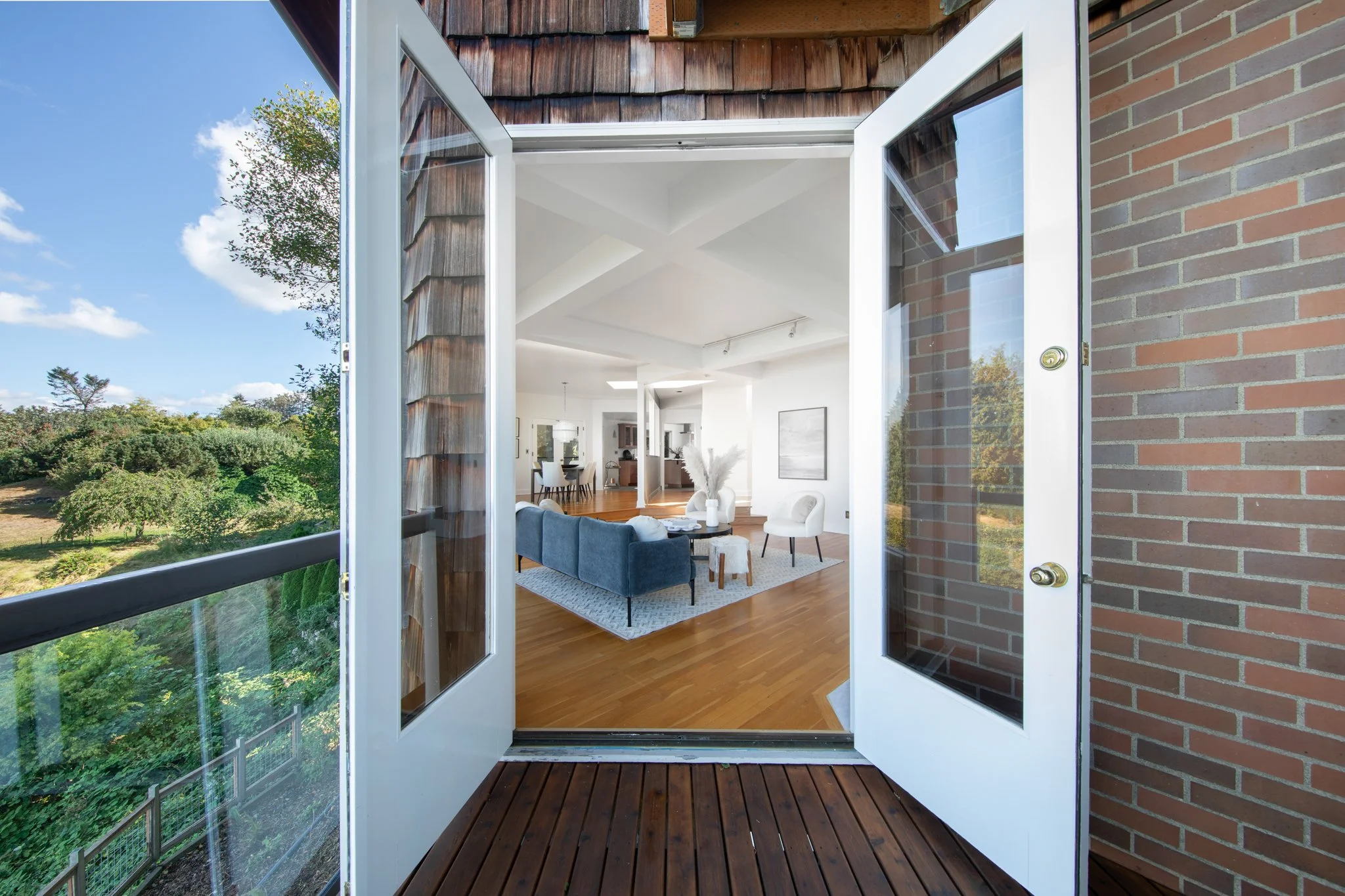 Open glass door leading to a living room with wooden flooring, white walls, and modern furniture, with a view of trees and blue sky outside.