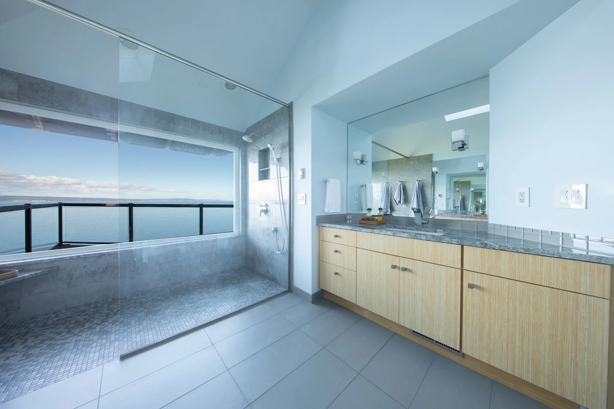 Modern bathroom with a walk-in shower featuring large glass windows overlooking a body of water, and a wooden vanity with a granite countertop.