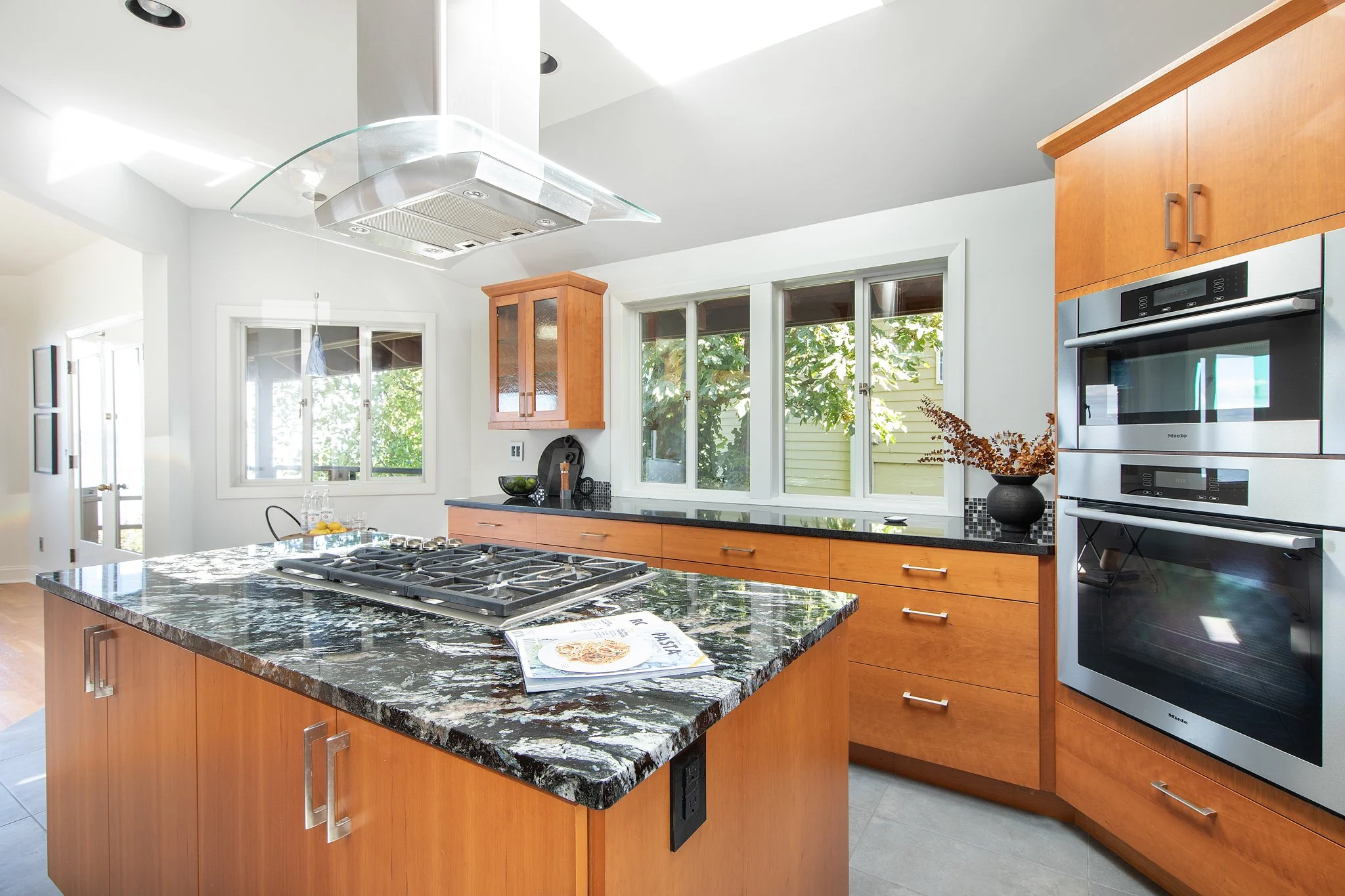 Modern kitchen with wooden cabinets, black granite countertops, stainless steel double oven, and a central island with a cooktop, large windows with a view of greenery, and sunlight brightening the space.