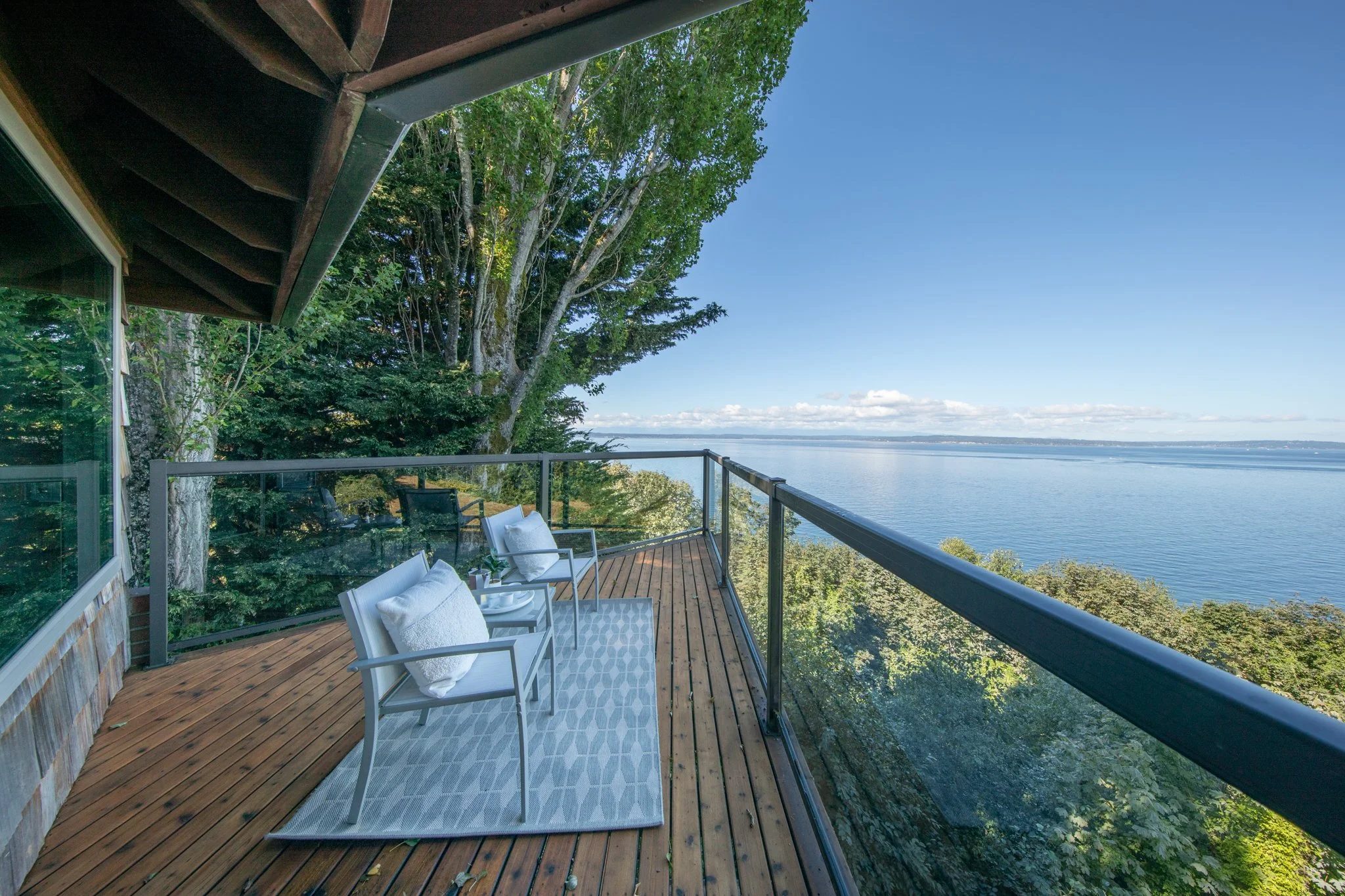 A wooden balcony with white chairs and towels, overlooking a large body of water with lush trees around, under a clear blue sky.