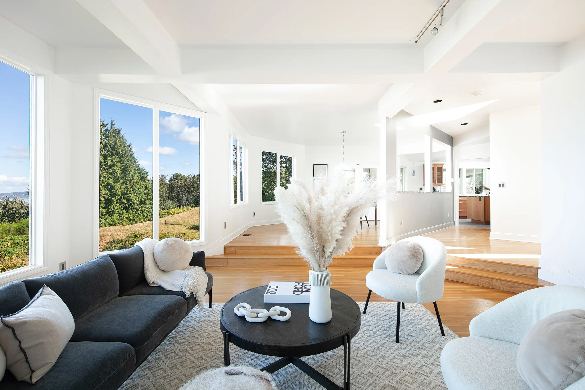 Modern living room with large windows, white walls, hardwood floors, black and white sofas, a black coffee table with decorative items, and a view of trees and sky outside.