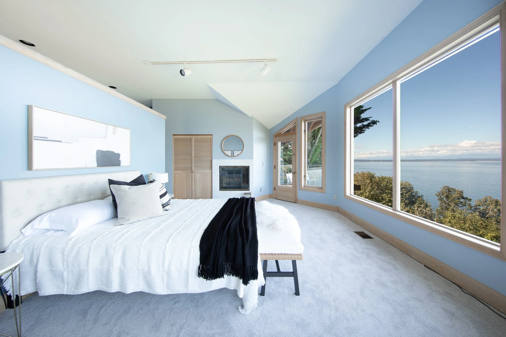 A bright bedroom with light blue walls and large windows overlooking a body of water and trees outside. The bed has white bedding, black and white pillows, and a black throw blanket. There is a small stool at the foot of the bed, a round mirror on th