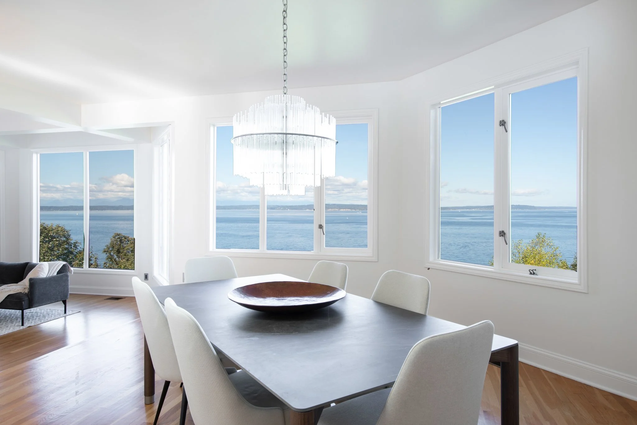 Interior view of a modern dining area with large windows showing a body of water and sky outside, a chandelier hanging from the ceiling, a round dining table with a decorative bowl, and chairs surrounding the table.