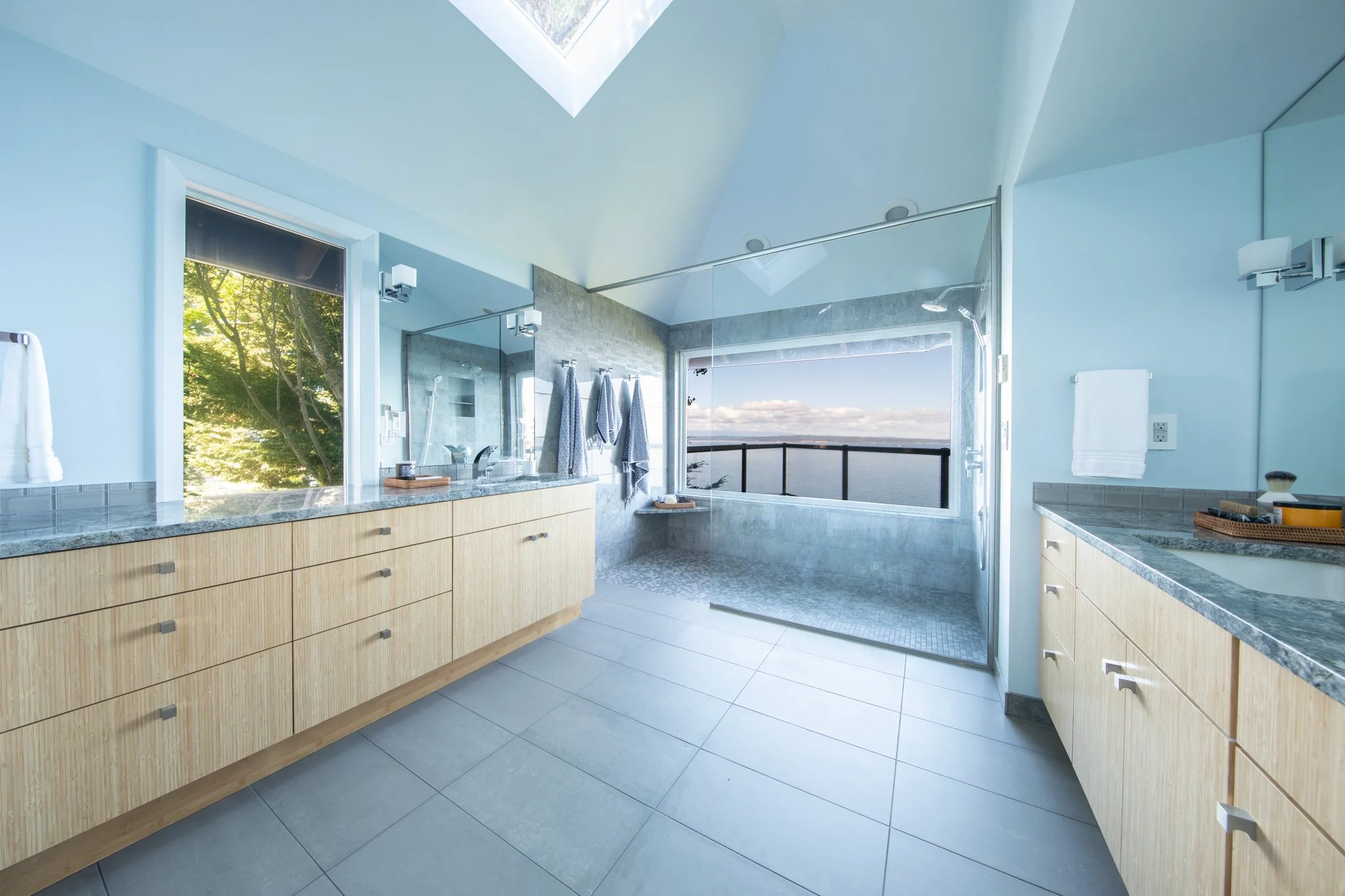 Modern bathroom with large window overlooking a water view, featuring light wood cabinetry, gray countertops, tiled shower, and towels hanging on the wall.
