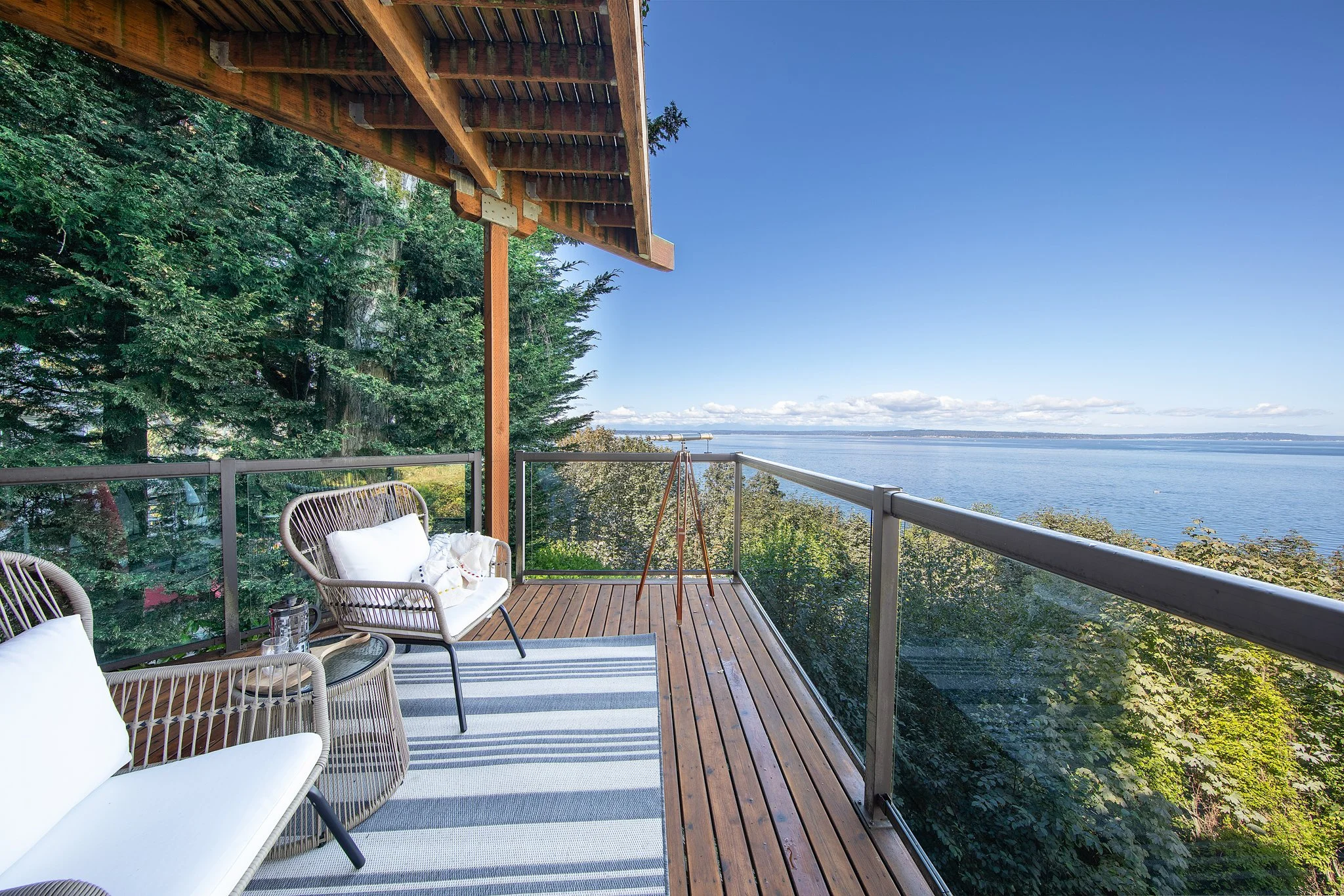 A balcony with outdoor furniture overlooking a large body of water under a clear blue sky, framed by lush green trees and a wooden overhang.