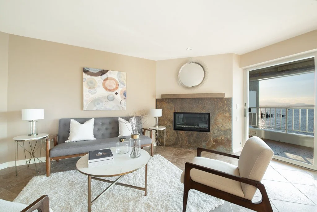 Living room with a gray couch, white pillow, glass coffee table, beige armchair, two white lamps, a fireplace with a circular mirror above, and sliding glass door leading to a balcony with ocean view.