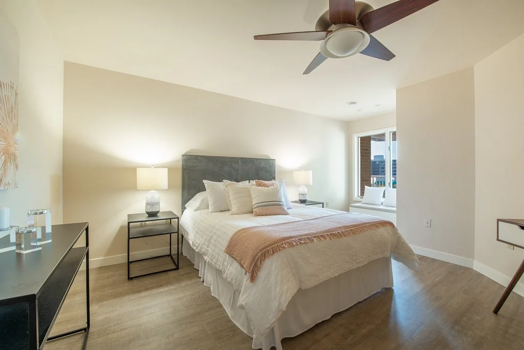 A bedroom with a bed, nightstands with lamps, a window with a bench, ceiling fan, and minimal decor.