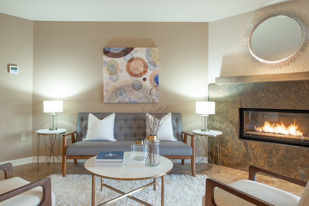 Living room with fireplace, art piece on beige wall, gray sofa with white pillows, round white coffee table with books and vases, two side tables with lamps, round mirror above fireplace.
