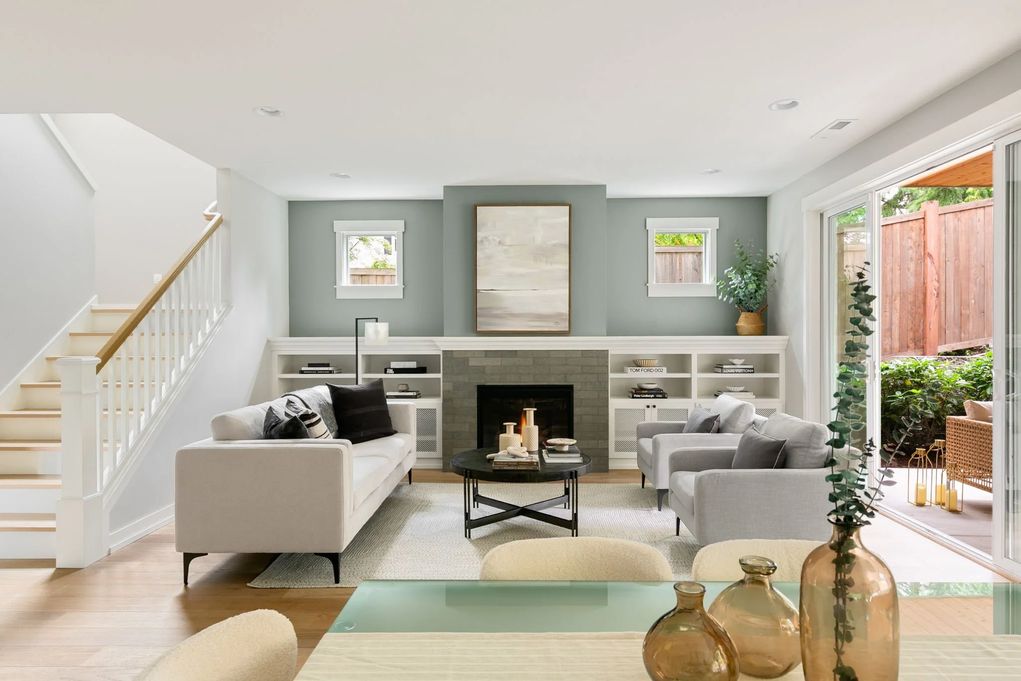 Living room with light gray walls, white trim, a fireplace, large windows, and sliding glass door opening to a fenced backyard. Furniture includes white sofas, a black coffee table, and greenery with decorative objects.