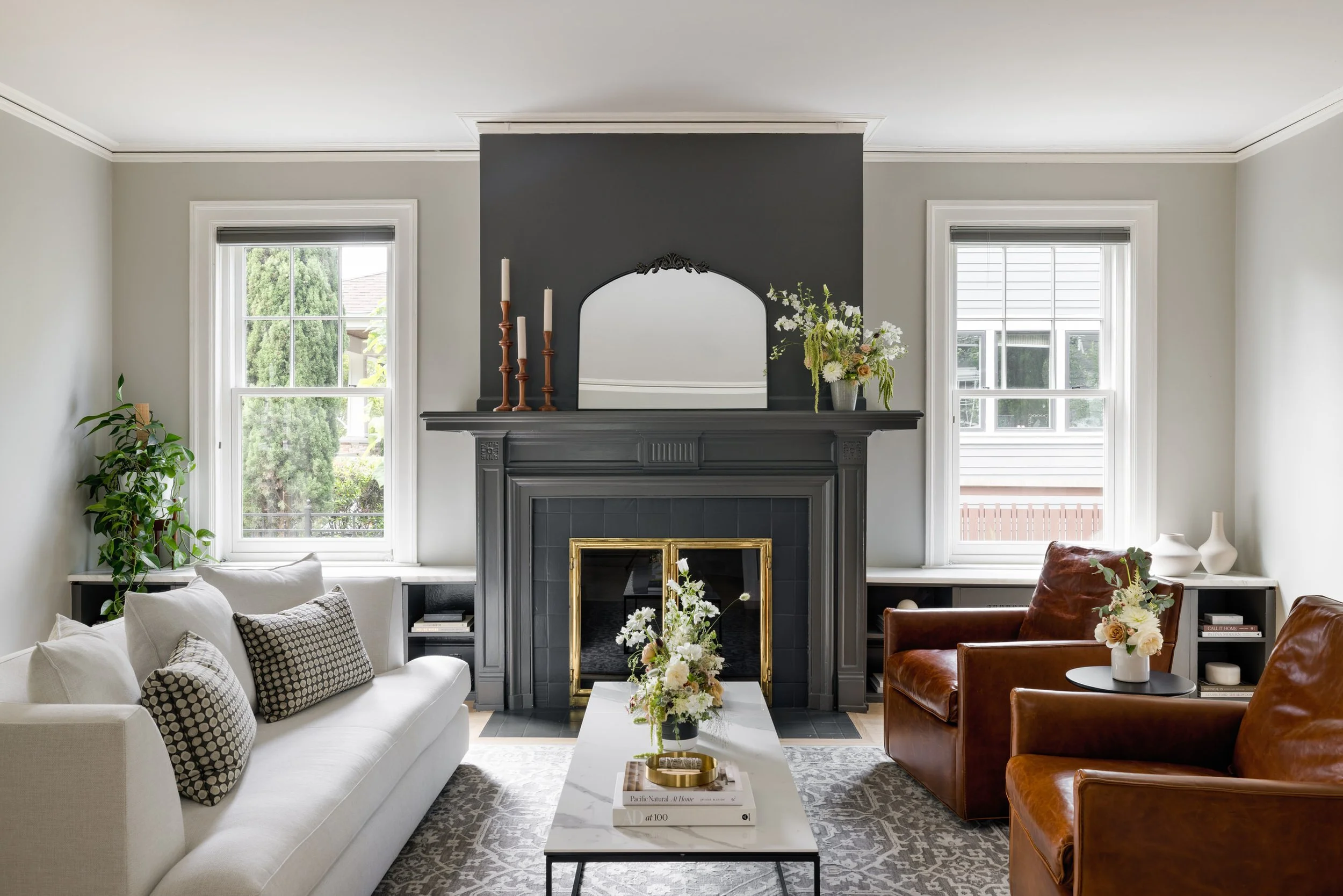 Living room with a gray fireplace, white sofa with patterned cushions, brown leather chairs, a white coffee table with books and a flower arrangement, two large windows, and decorative items on the mantle and side tables.