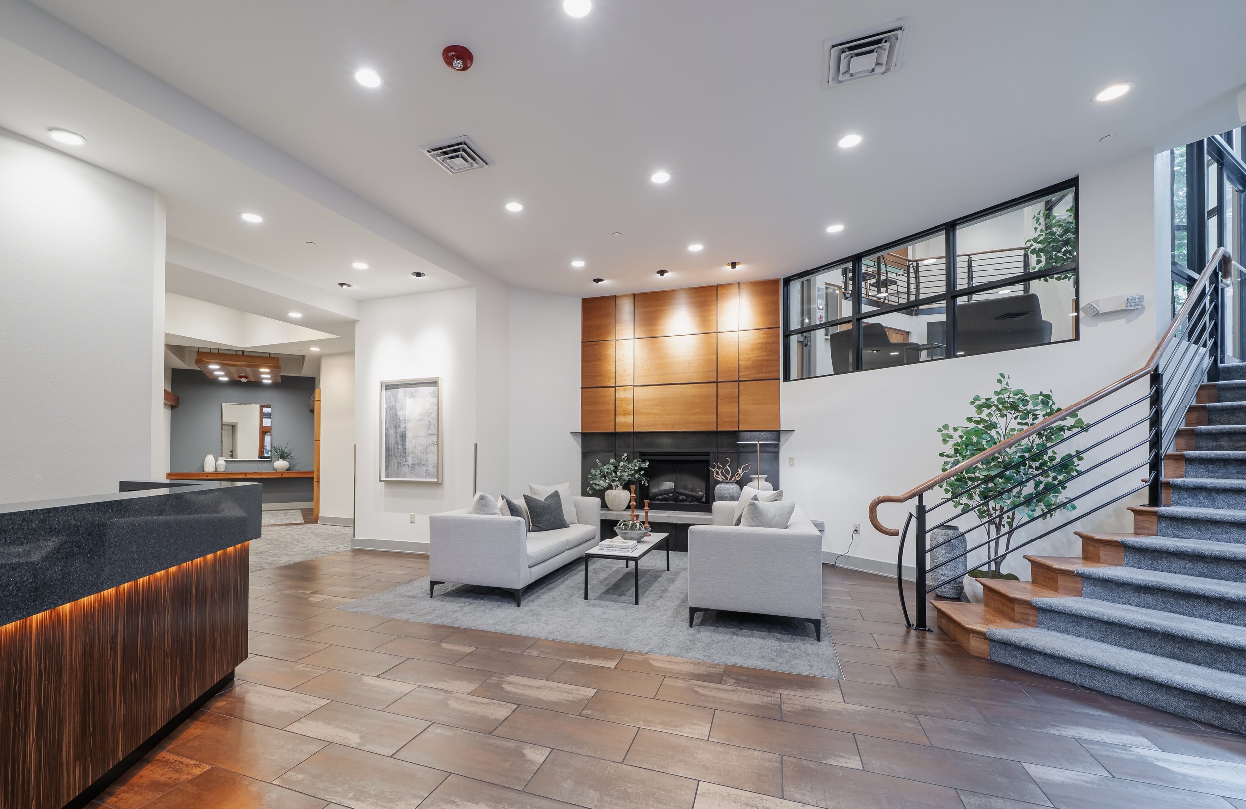 Interior of a modern, well-lit lobby or lounge area with white walls, wooden accents, a fireplace, beige sofas, a glass staircase with metal handrails, and large windows letting in natural light.
