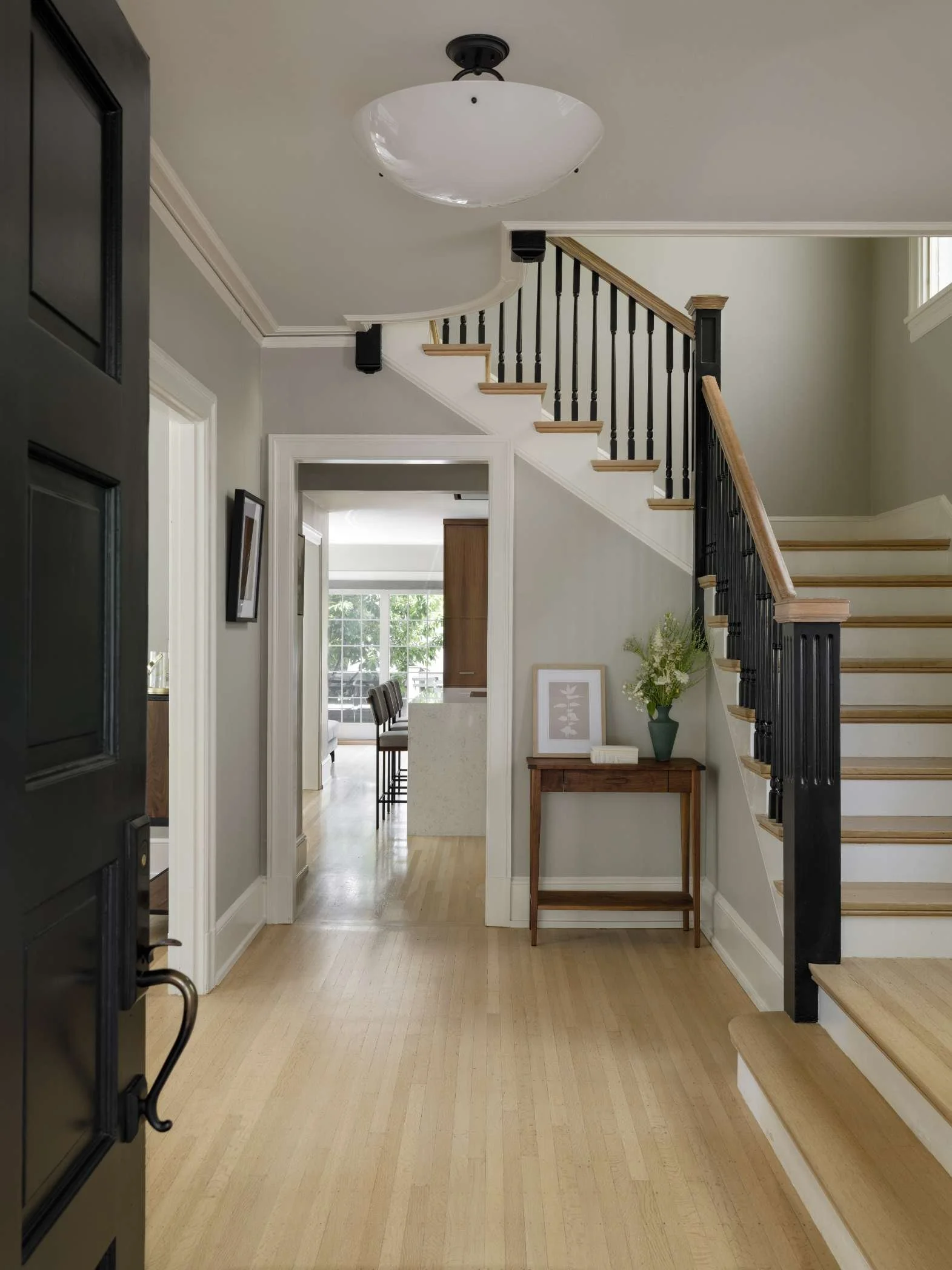 Interior of a home showing a staircase with black and wood banister, a hallway with a light wood floor, and a view into a dining room with a table and chairs, with a window and greenery outside.