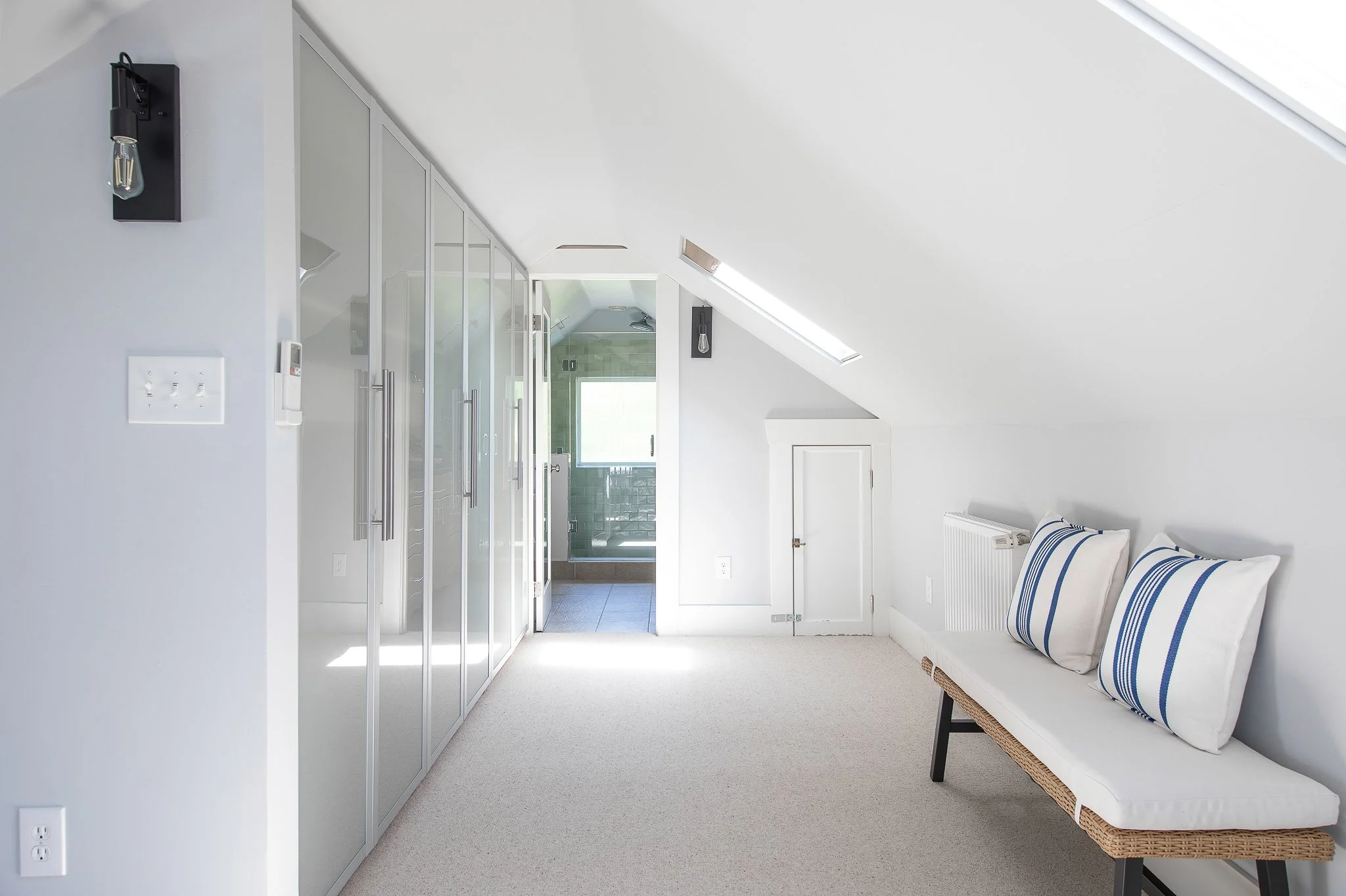 A bright, minimalist attic bedroom with white walls and sloped ceiling. Features a bench with striped cushions, a closed white door, glass-fronted closet, and a skylight above showing a view of the sky. A radiator is visible next to the bench.