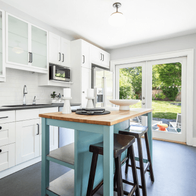 Kitchen with white cabinets, a wooden center island painted light blue, a stainless steel refrigerator, microwave, glowing natural light from sliding glass door, and view of green backyard.