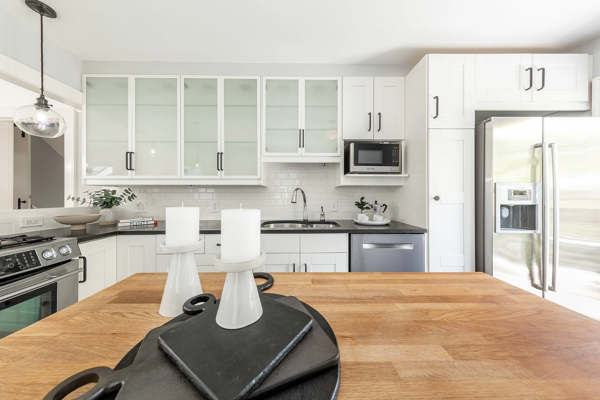 Modern white kitchen with upper cabinets, black handles, a microwave, a stainless steel refrigerator, and decorative plants on the counter.