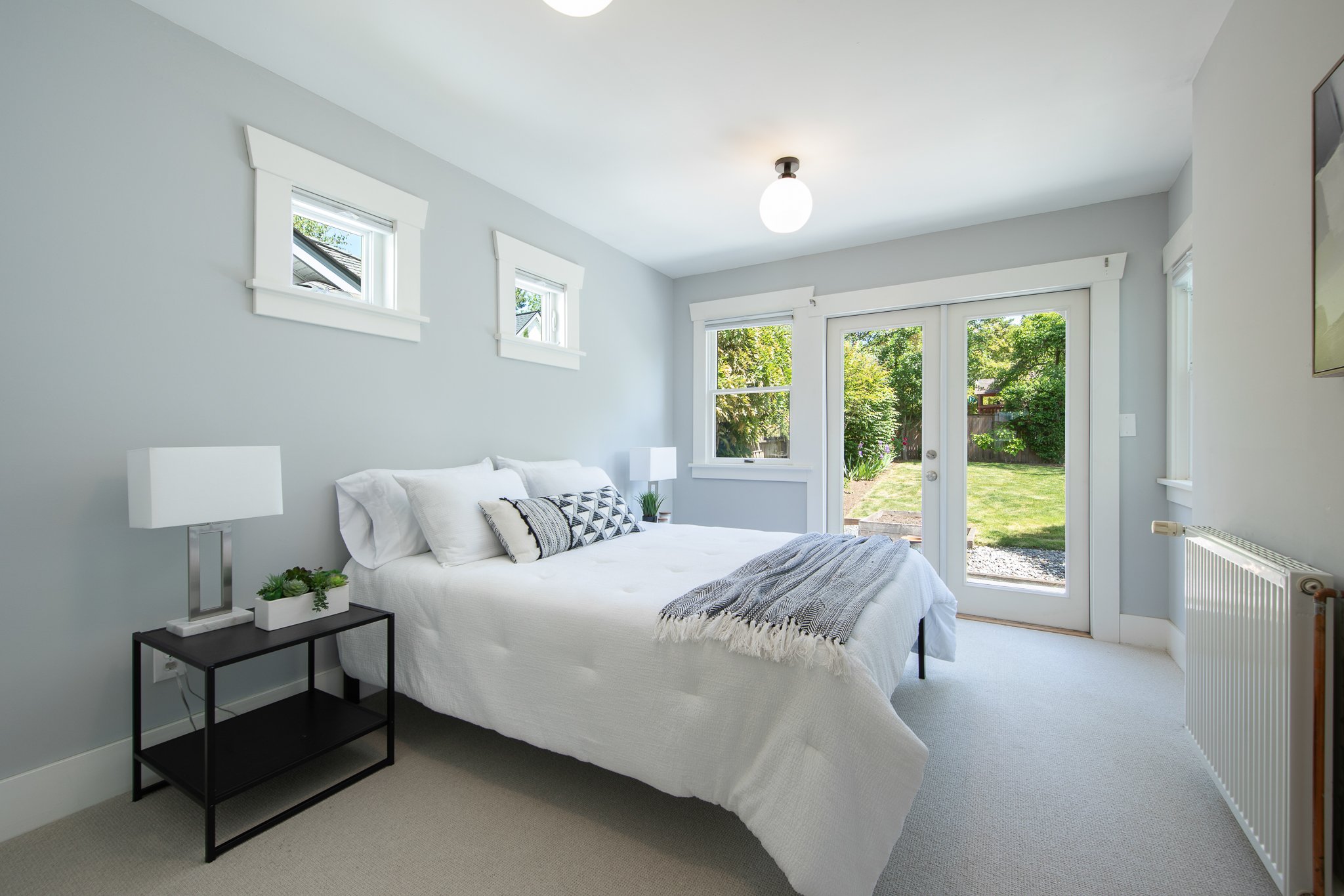 A bright bedroom with a large window and glass door overlooking a garden. The bed has white bedding and patterned pillows, with a nightstand and lamps on either side. White trim decorates the light gray walls.