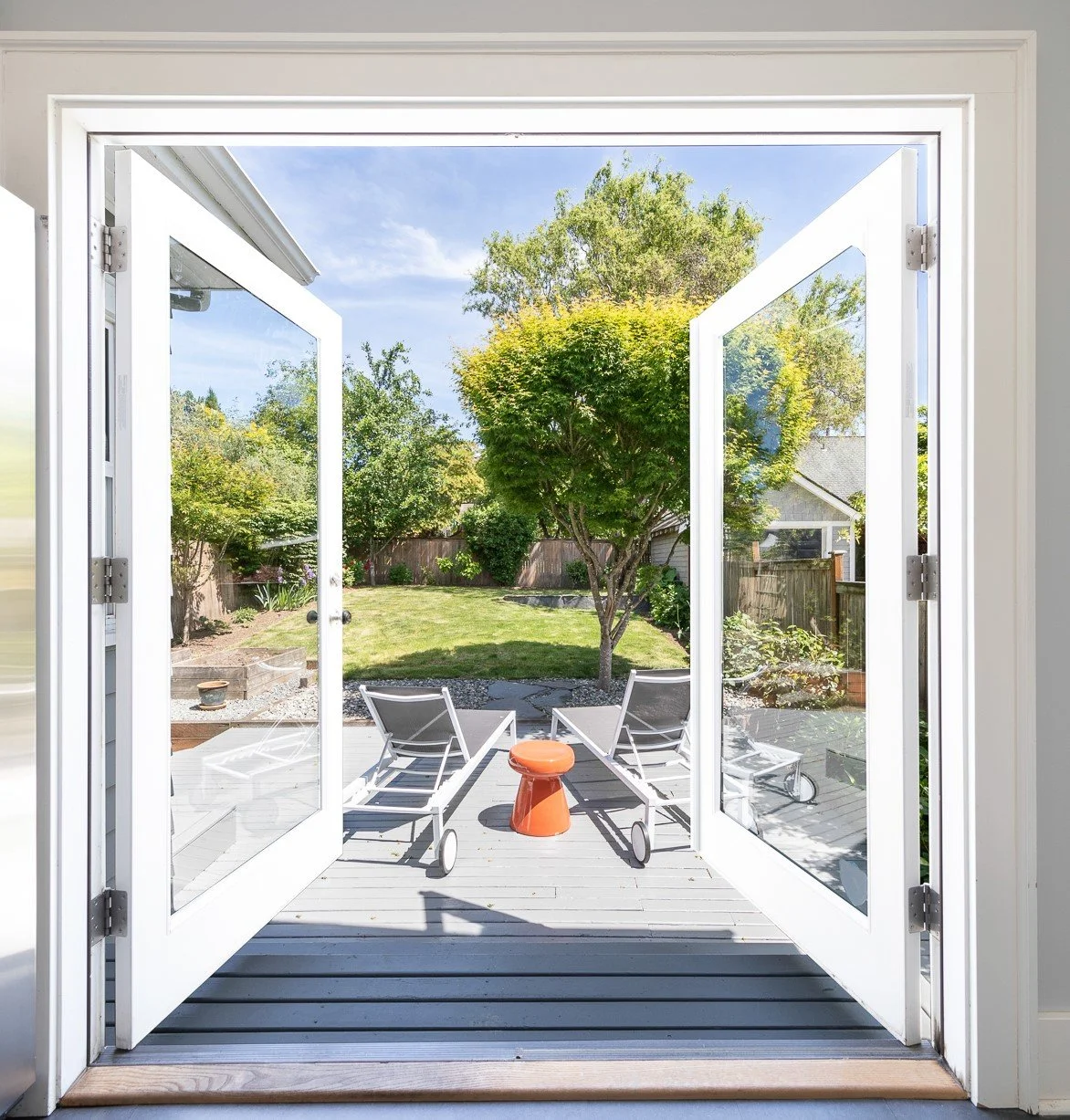 Open white French doors leading to a backyard with green lawn, trees, garden, and two lounge chairs with a small orange table between them.