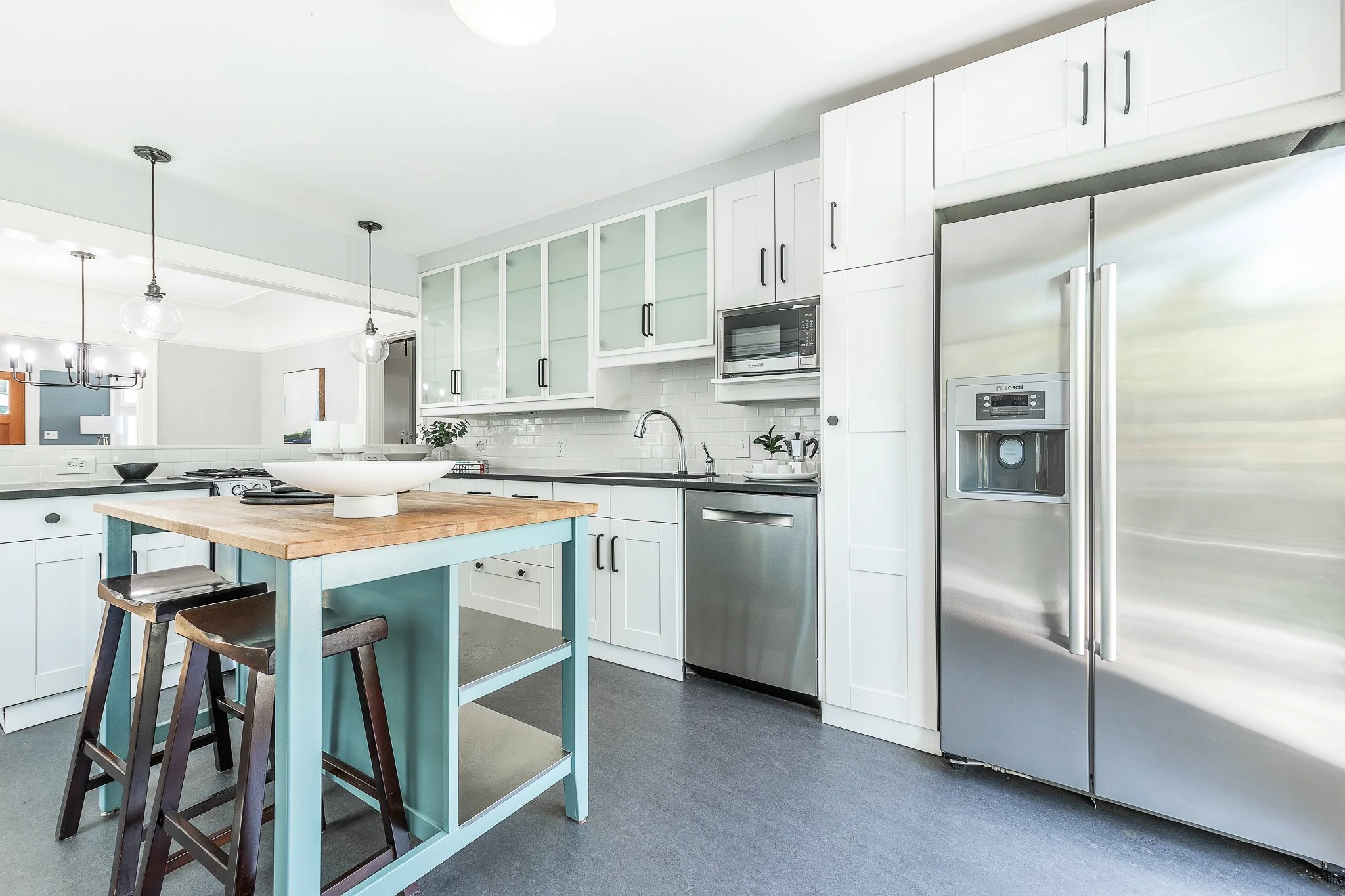 Modern white kitchen with stainless steel refrigerator, microwave, and cabinetry, island with wooden countertop, and pendant lights.