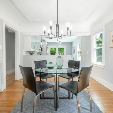 Dining room with a glass table, six black chairs, a modern chandelier, and windows overlooking a yard.
