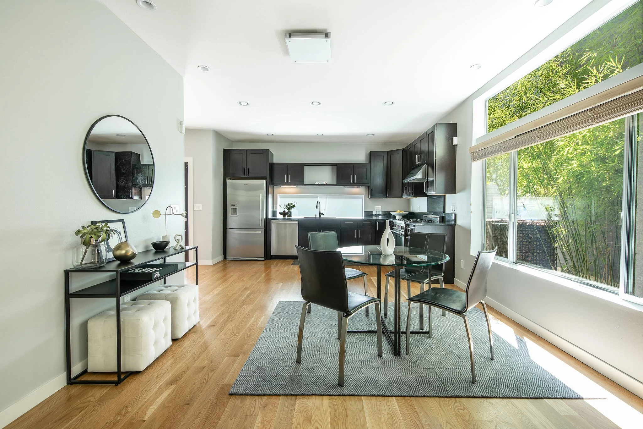 A modern kitchen and dining area with black cabinets, stainless steel refrigerator, large windows with blinds, a round glass dining table with black chairs, a black console table with decor, and a light-colored bench with tufted seats.
