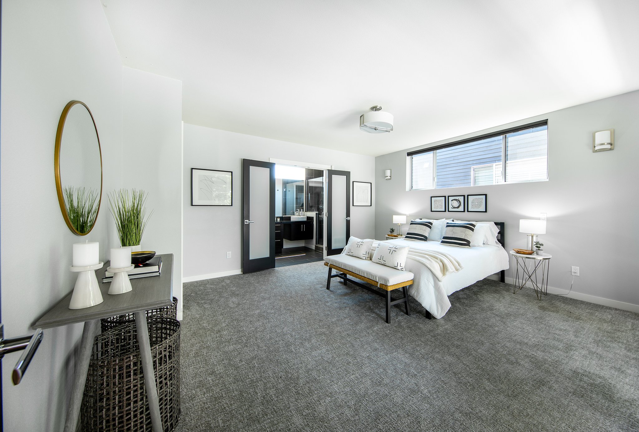 A modern bedroom with a white bed, black headboard, and black and white striped pillows. Art prints hang above the bed, and there are white lamps on nightstands. A small bench sits at the foot of the bed. A window with a view of another building is a