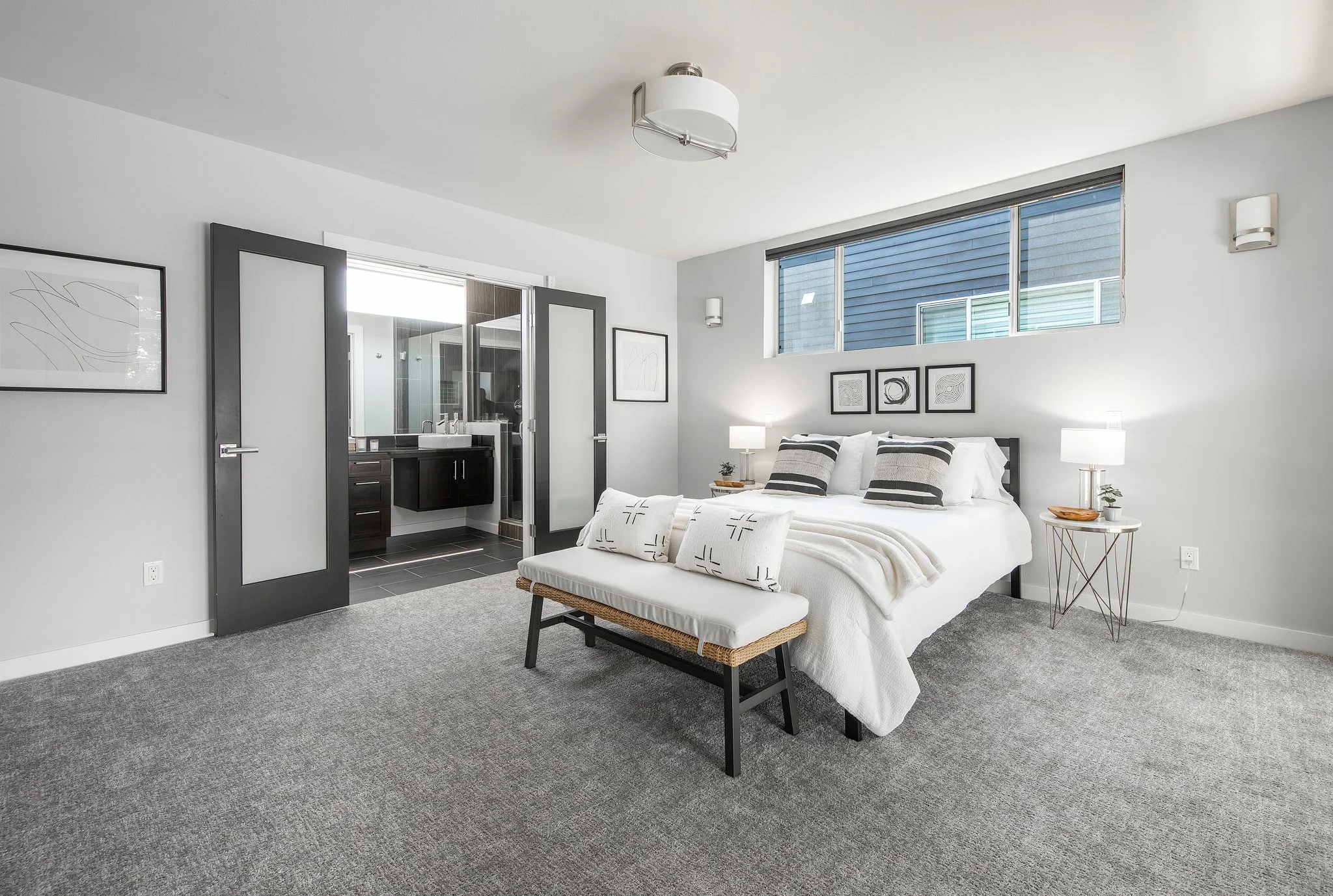 A contemporary bedroom with white walls, a bed with black and white pillows, nightstands with lamps, abstract wall art, and a window with blinds. An open door reveals a bathroom with a sink and mirror.