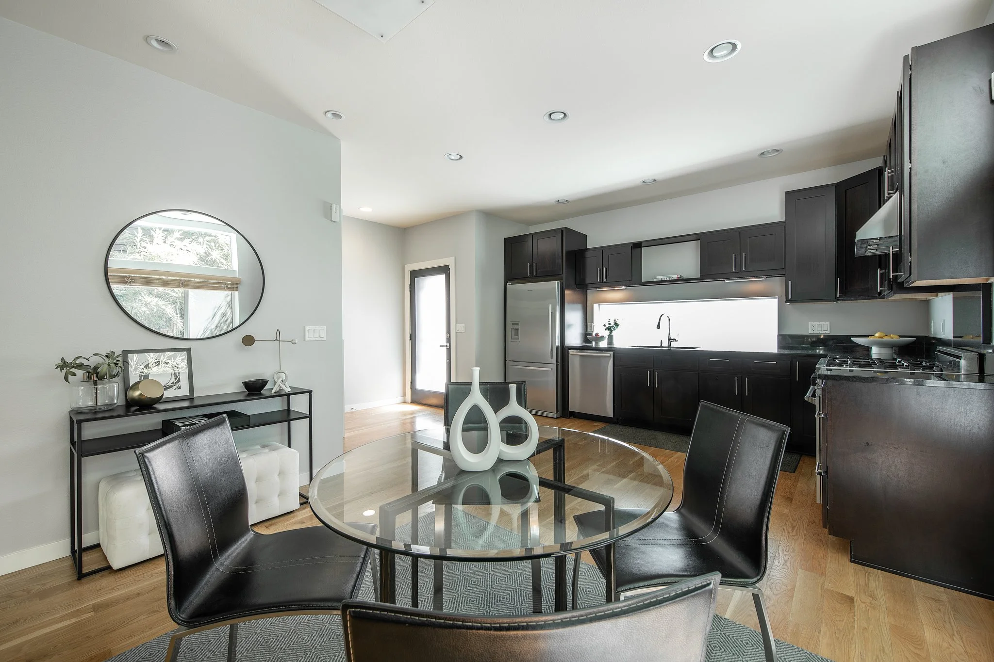 Modern kitchen and dining area with black cabinets, stainless steel appliances, a glass dining table with black chairs, and decorative white vases on the table. A round mirror and decor are on the wall.