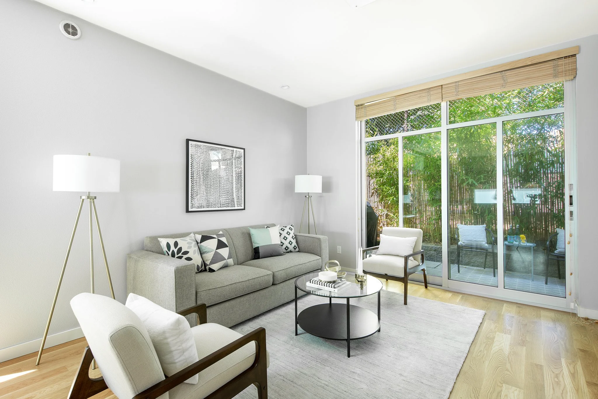 Living room with gray sofa, white armchair, black round coffee table, two white floor lamps, wall art, large window with bamboo blinds, and a view of an outdoor patio with chairs and greenery.