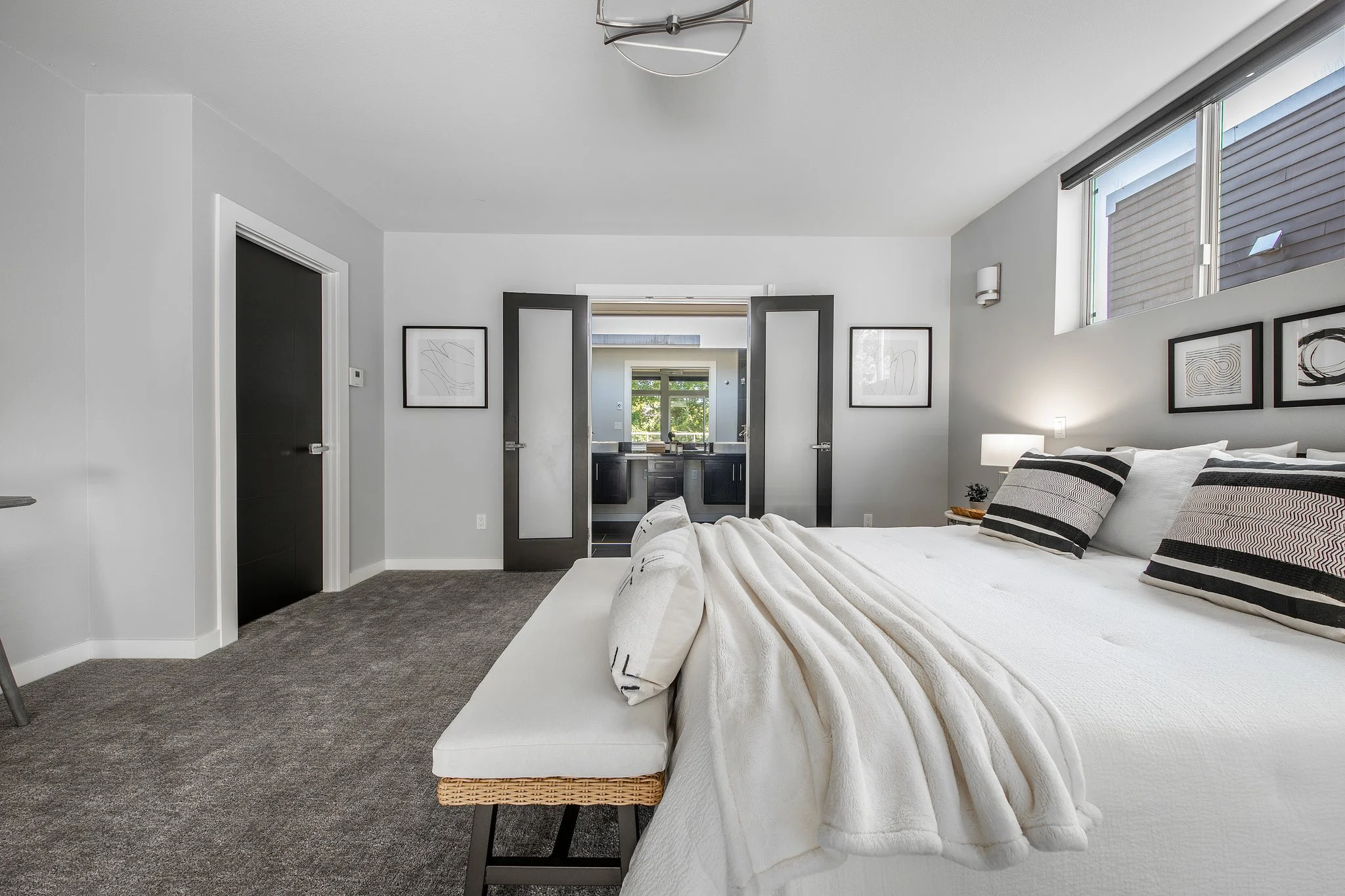 A modern bedroom with a white bed, black and white striped pillows, and a white quilt. The room has gray carpet, white walls with minimalistic art, and two black doors leading to a bathroom with a double sink. There are large windows with gray blinds