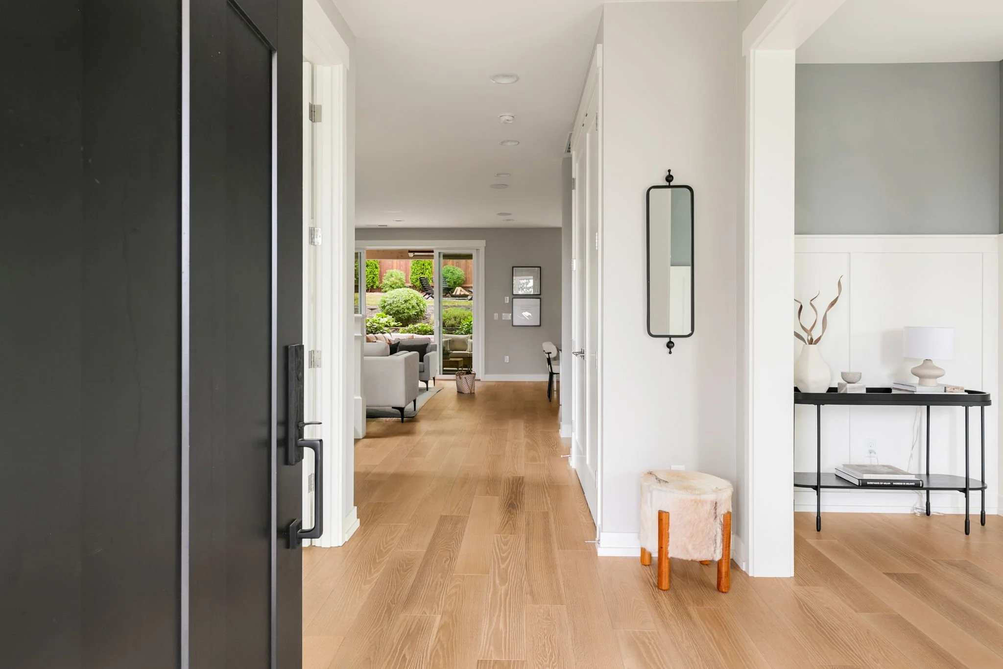 View from inside the hallway of a modern home, looking into the living room and backyard, with wooden floors, white walls, and contemporary decor.